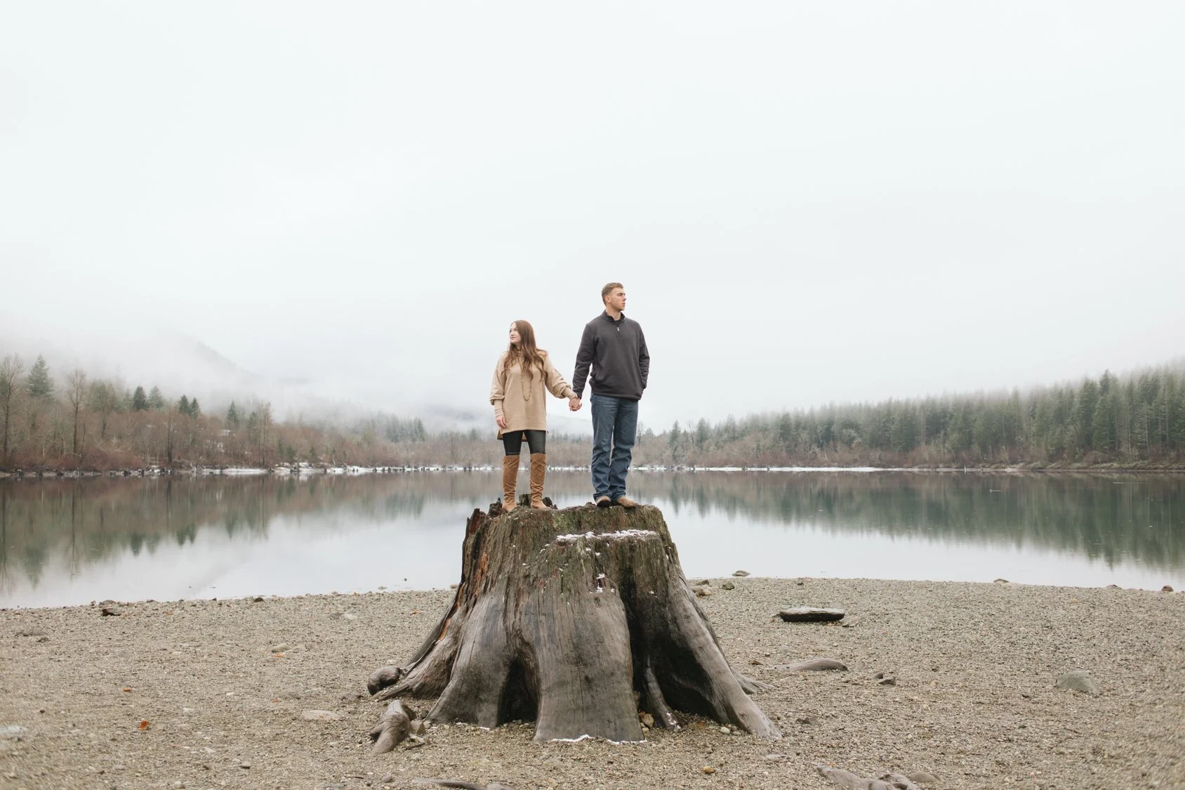 Two people, a woman and a man, holding hands and standing on a large tree stump by a calm lake with a forested and foggy backdrop.