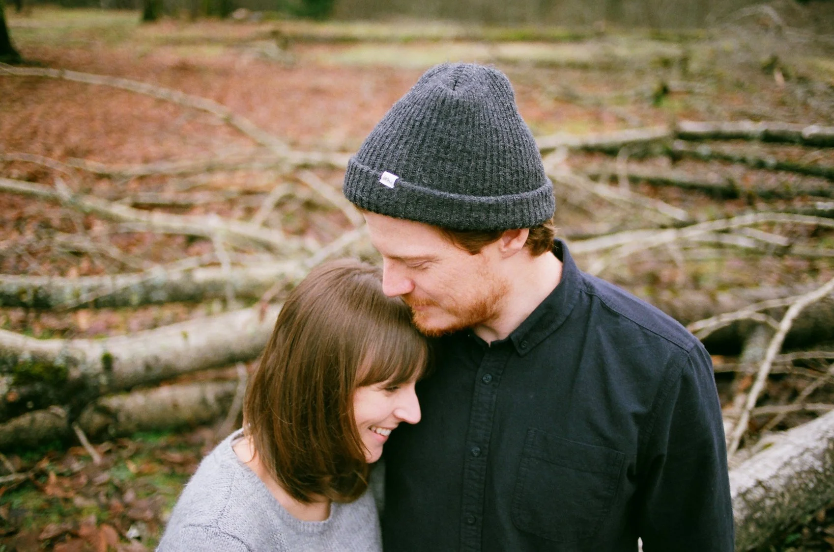 A man and woman sharing a hug outdoors in a wooded area, with fallen logs and branches in the background.