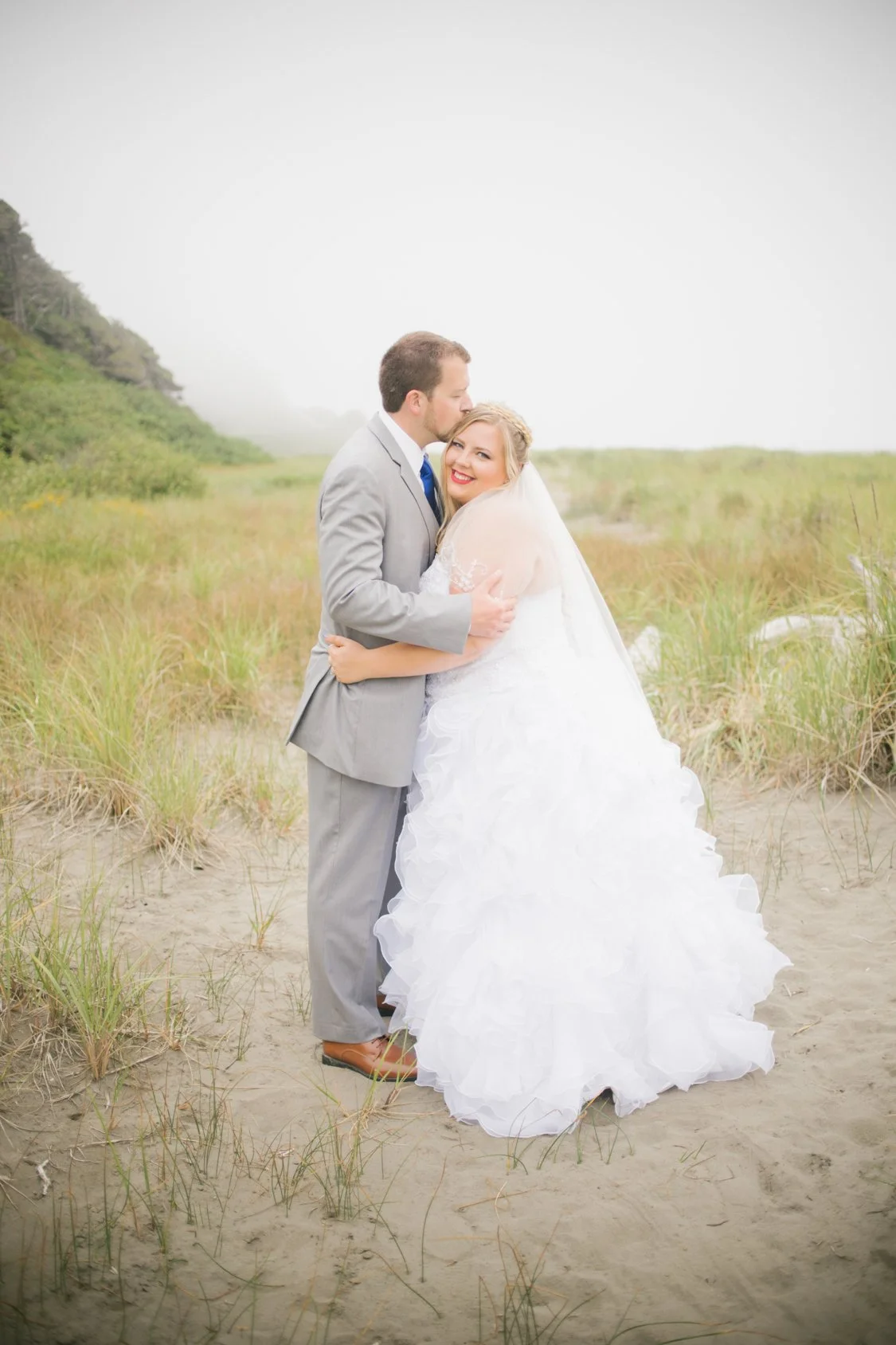 A newlywed couple in wedding attire embracing in a grassy, sandy outdoor area with a hill and trees in the background.