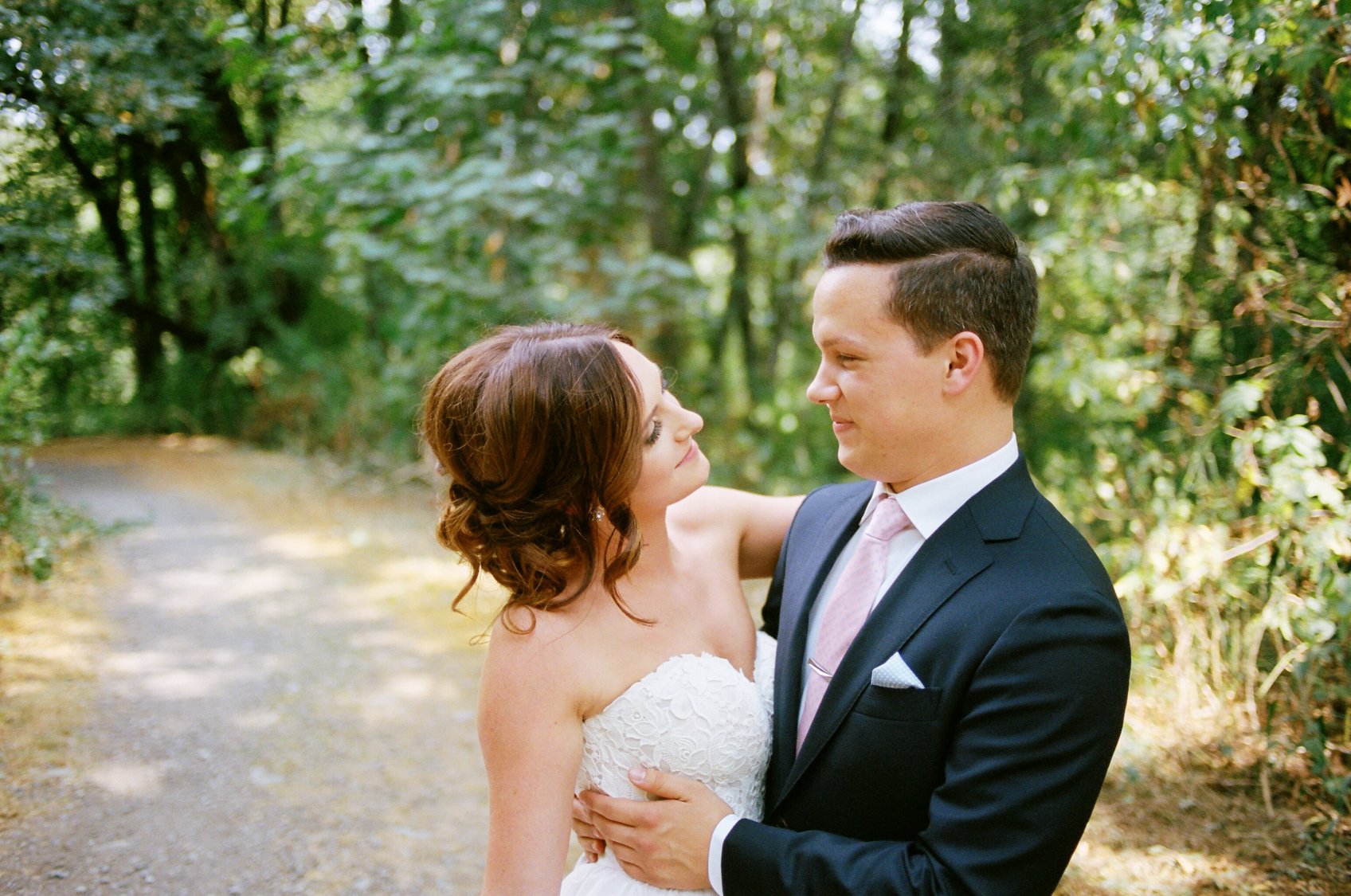 A bride and groom holding each other in a forest. The bride is wearing a white strapless wedding dress and the groom is in a dark suit with a light pink tie.