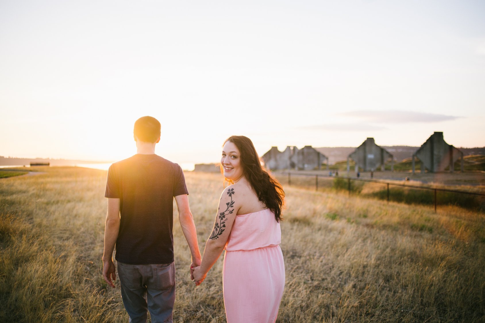A smiling woman with long dark hair and a tattoo on her arm holds hands with a man with short hair, walking through a grassy field during sunset with old industrial buildings in the background.