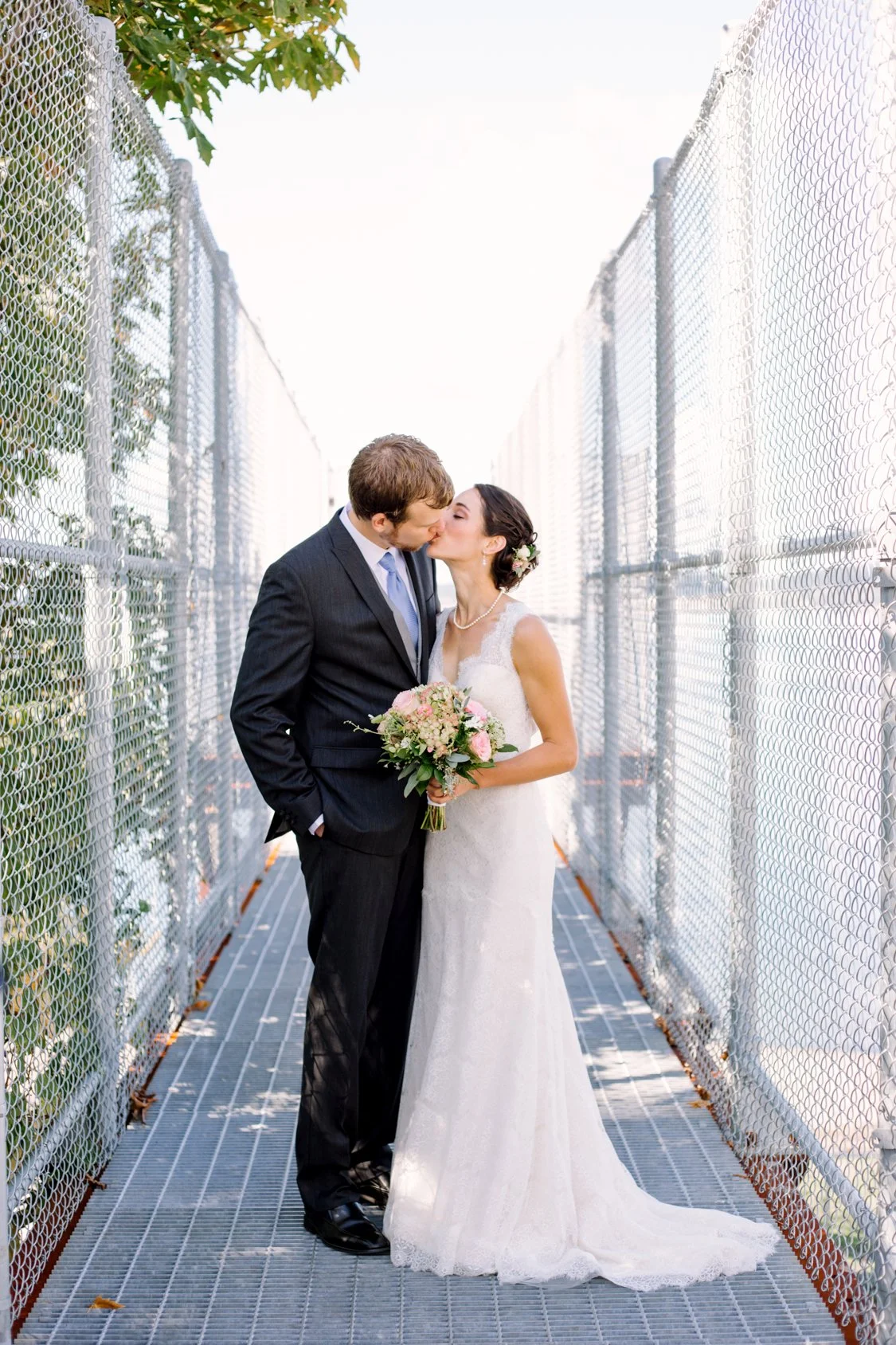 A bride and groom share a kiss, standing in a narrow outdoor corridor with metal fencing on both sides. The bride is holding a bouquet of pink and white flowers, wearing a white wedding dress, and has dark hair adorned with a floral accessory. The gr