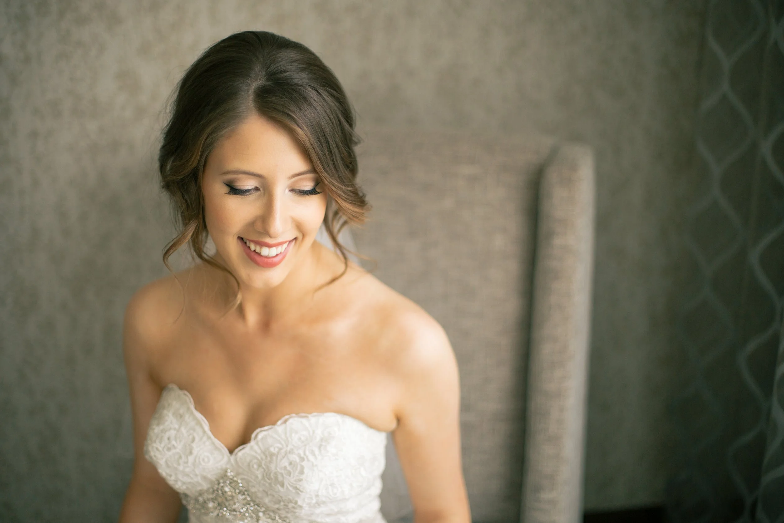 A woman with shoulder-length brown hair, styled in loose curls, wearing a white lace dress, smiling and looking down, against a textured neutral background.