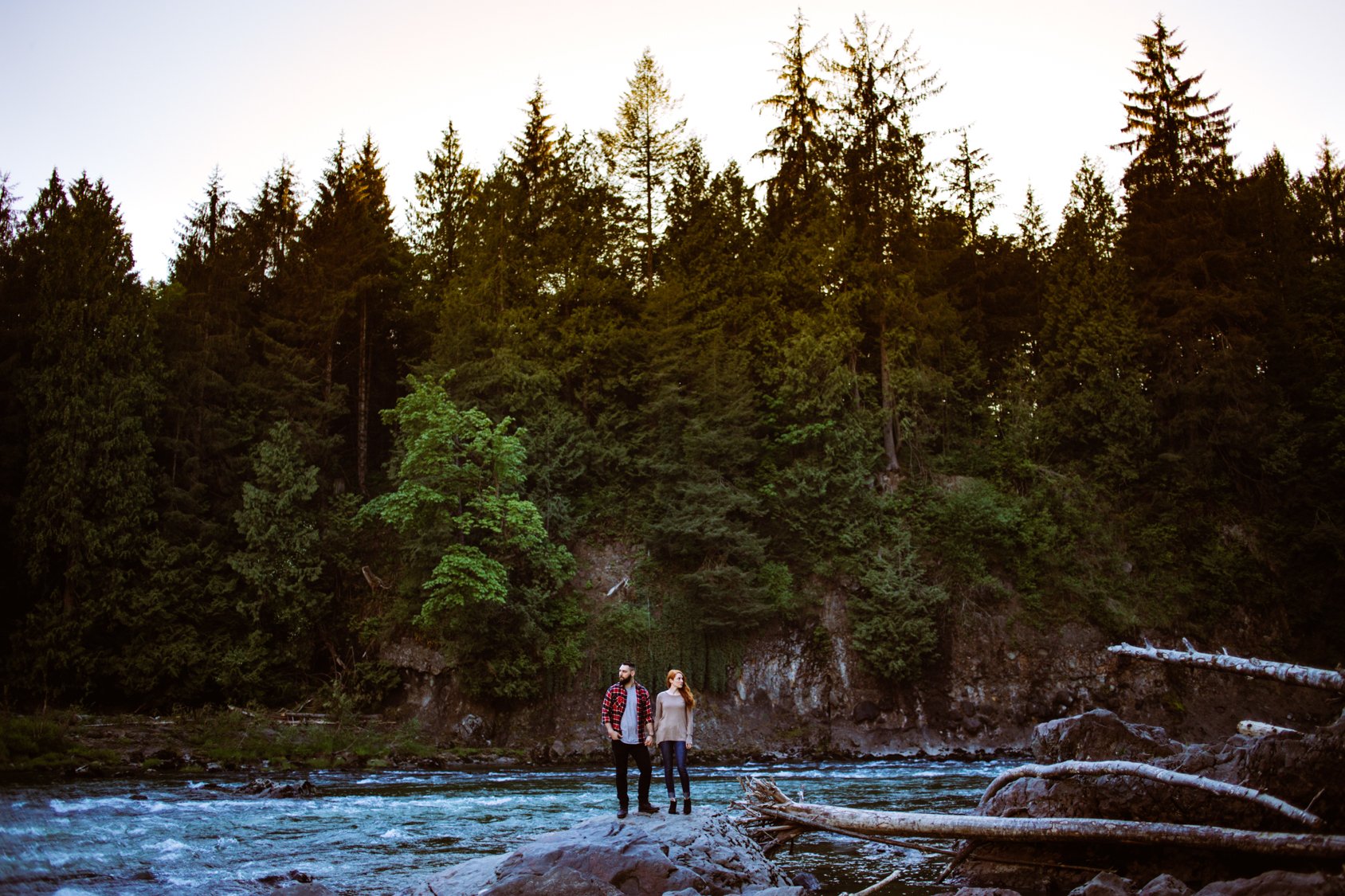A man and woman holding hands and standing on a rock in a flowing river, with a dense forest of tall green trees in the background.