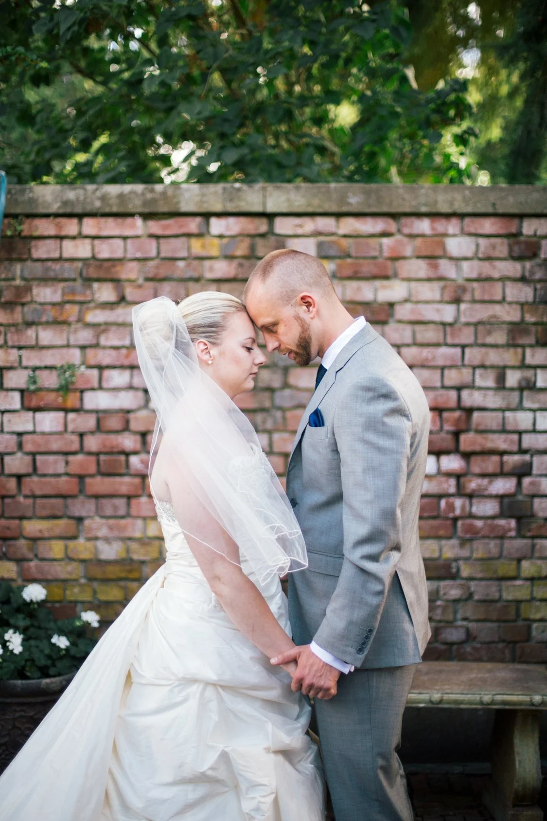 Bride and groom standing with foreheads touching, holding hands, outdoors in front of a brick wall, with greenery in background.