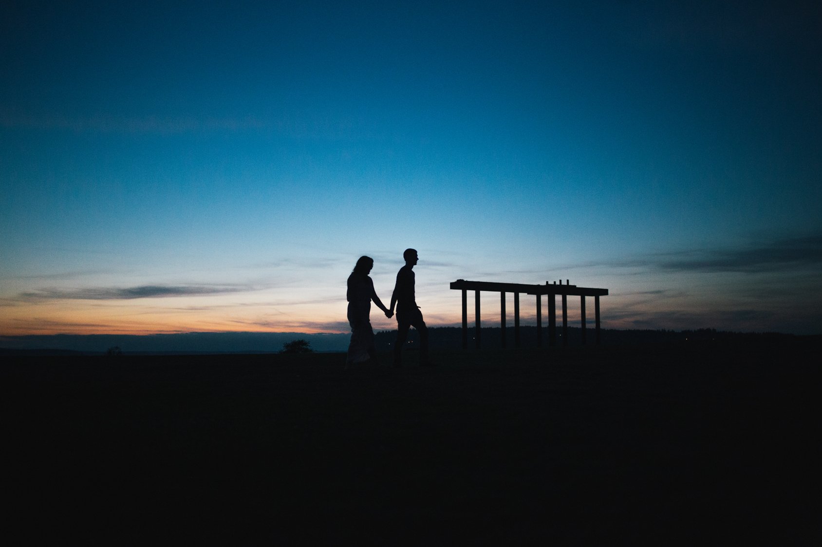 Silhouettes of a man and woman holding hands walking outdoors at dusk, with a wooden platform or structure and a colorful sky in the background.