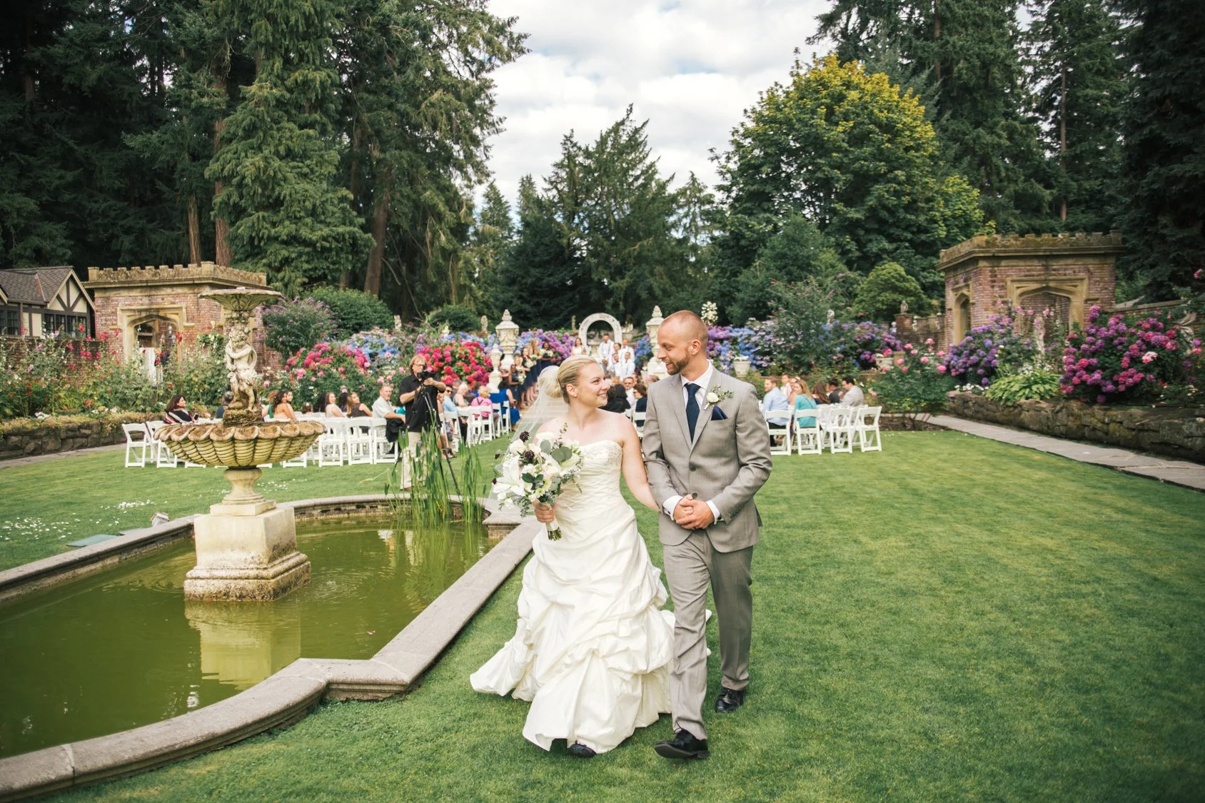 A bride and groom walking hand-in-hand in a garden during their wedding reception, with guests seated at tables in the background and surrounded by trees and flowers.
