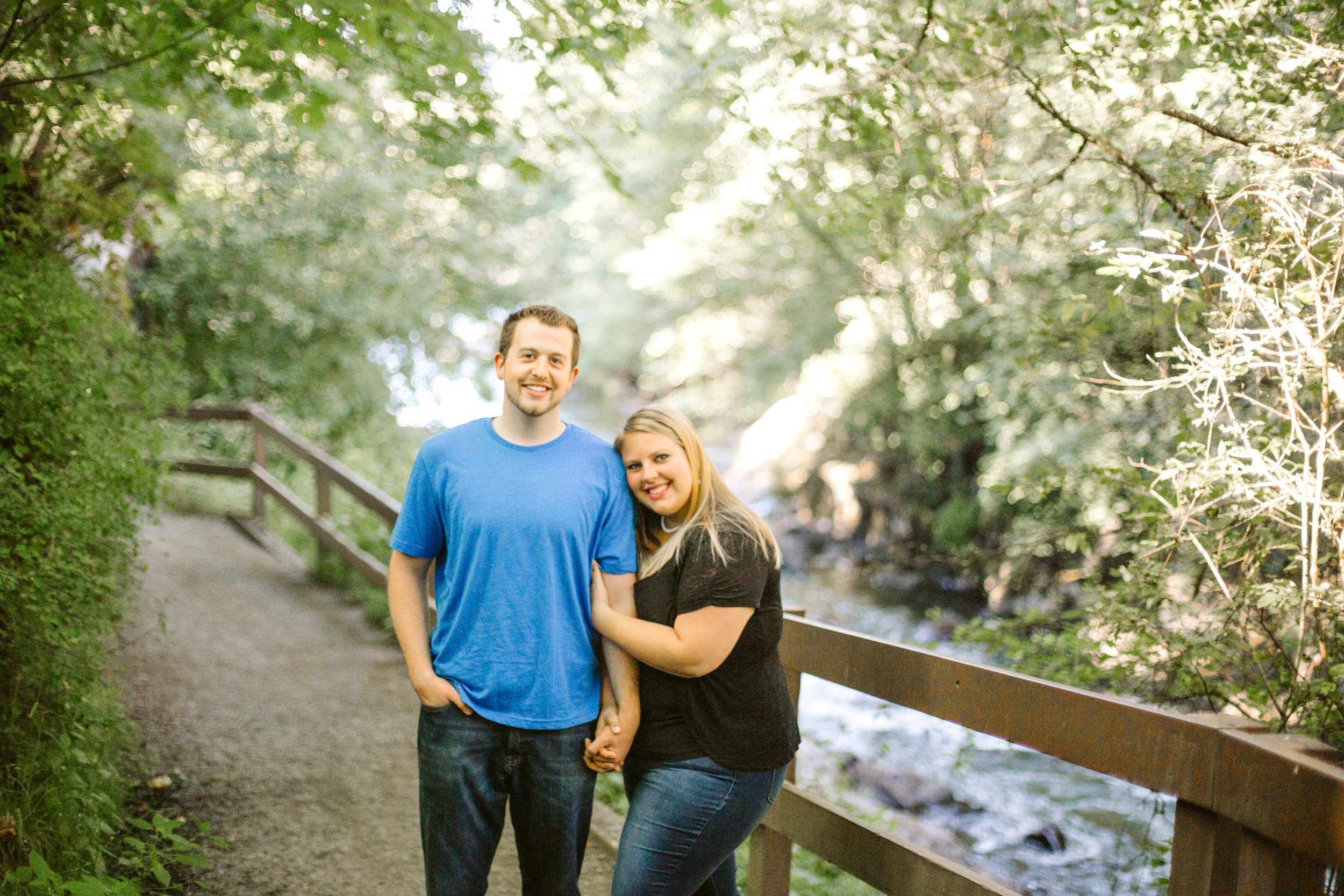 A smiling man and woman holding hands, standing on a wooded path near a railing over a river.