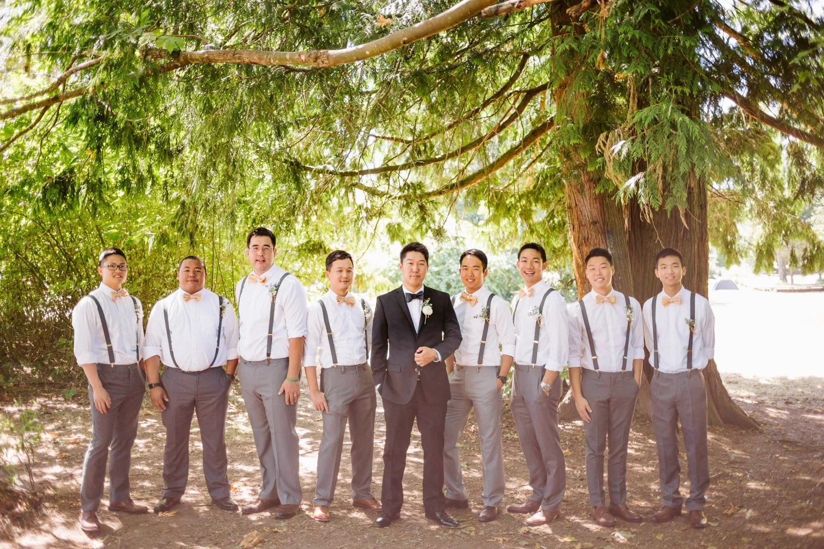 Group of men in formal attire standing outdoors in front of a large tree, dressed for a wedding, with the groom in the center wearing a black tuxedo and bowtie.
