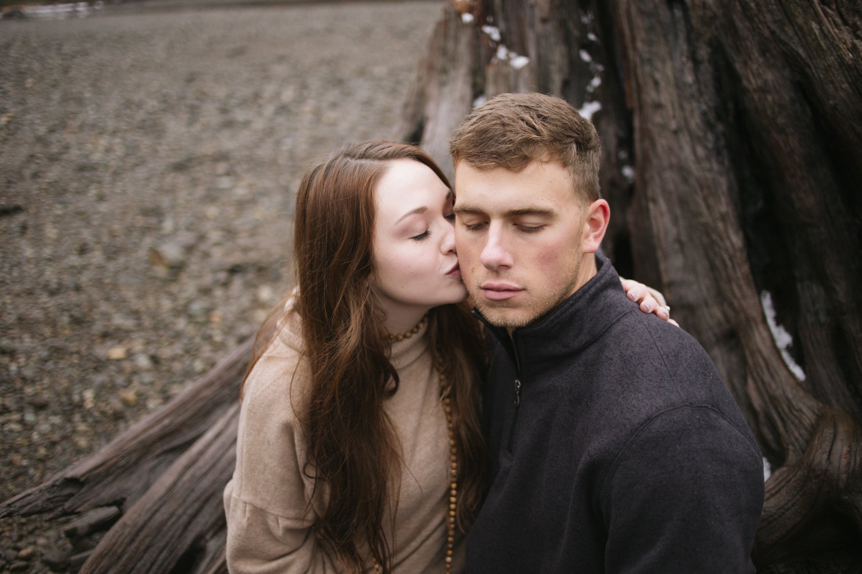 A young woman with long brown hair giving a kiss on the cheek to a young man with short light brown hair who has his eyes closed, standing next to a large piece of driftwood on a rocky beach.