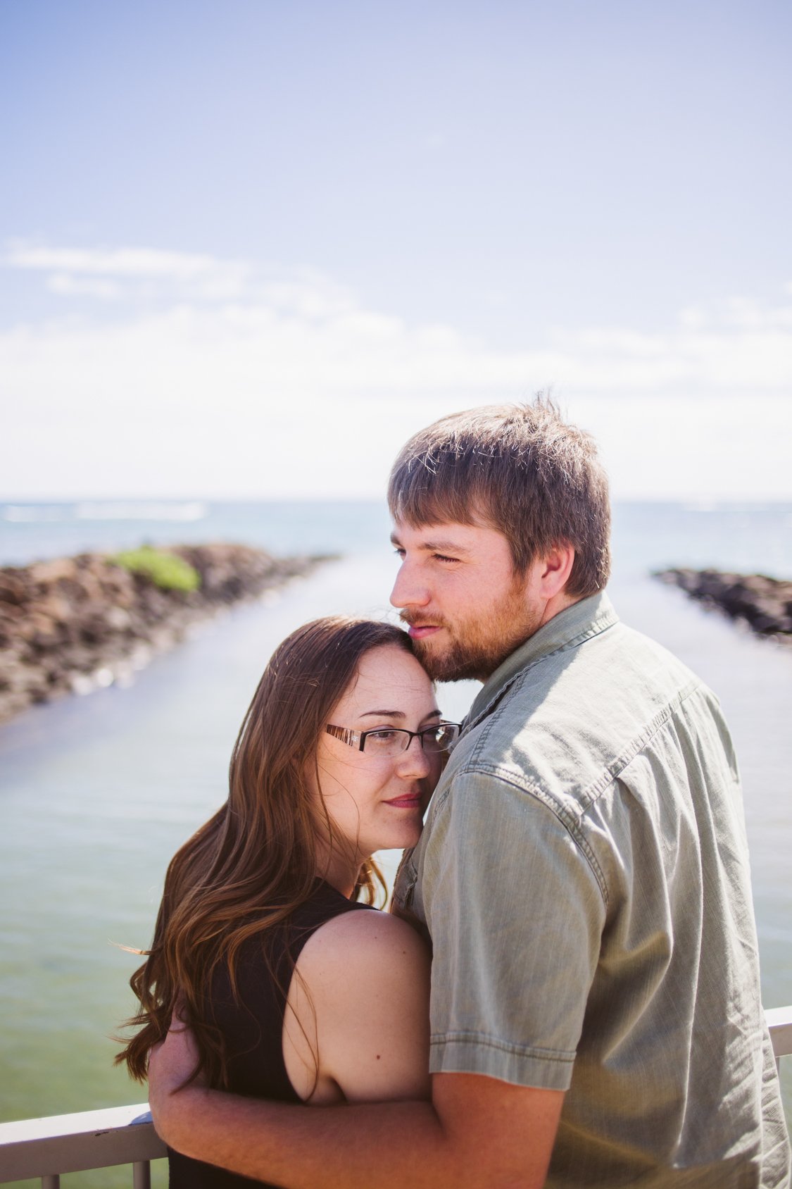 A young woman and man embrace outdoors near water, with rocky shores in the background on a sunny day.