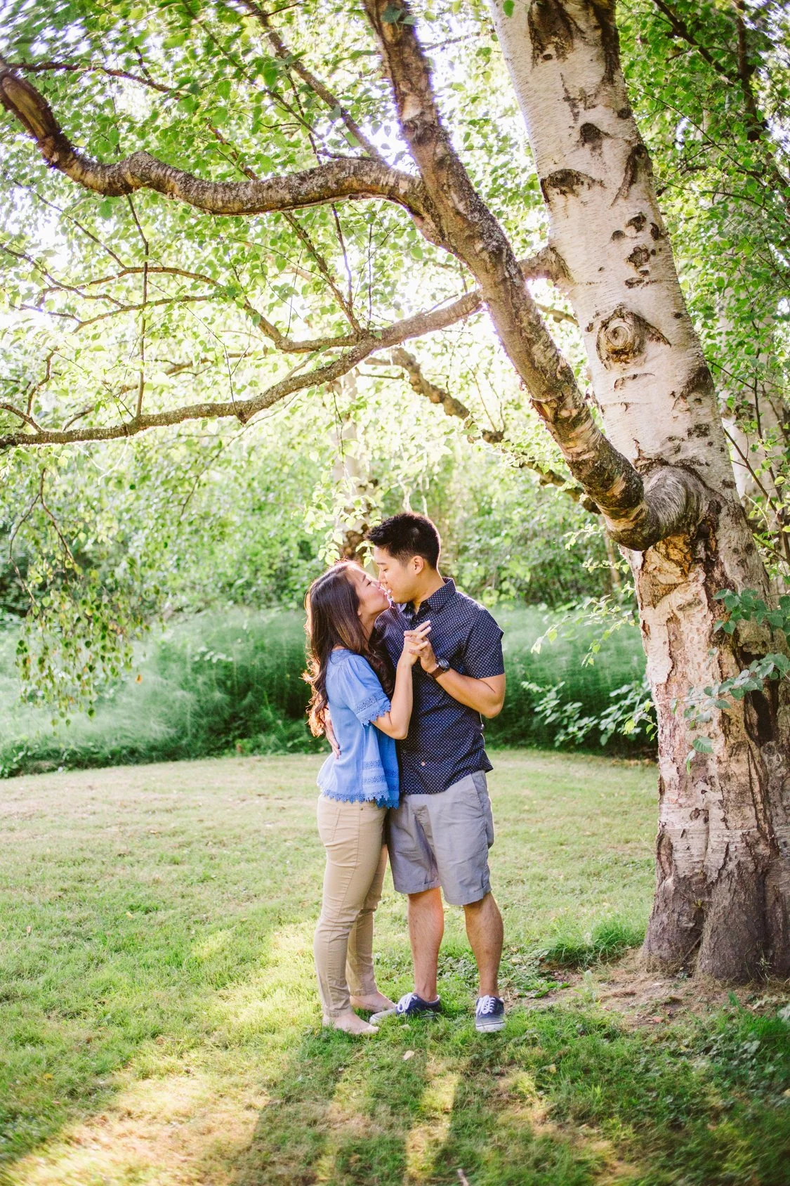 A young couple is sharing a kiss outdoors under a large tree with lush green leaves. The woman has long dark hair, wearing a blue top and beige pants. The man has short dark hair, wearing a dark blue short-sleeved shirt and gray shorts. They are embr