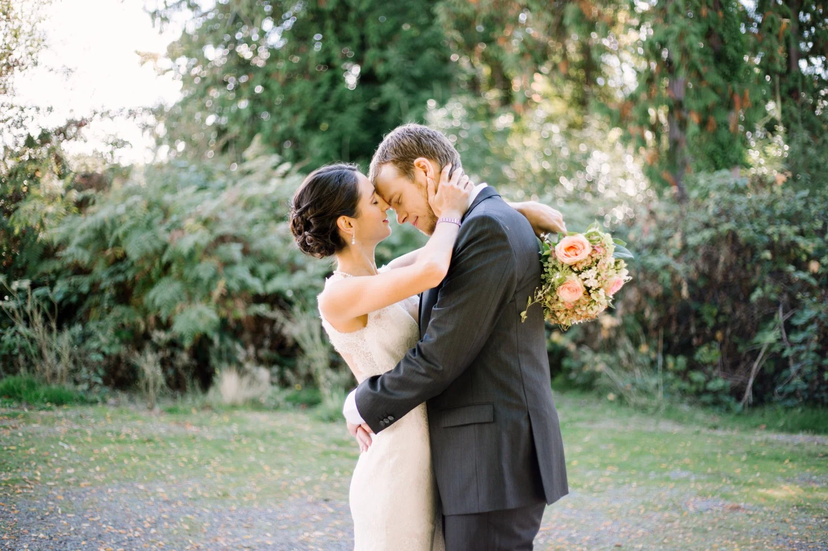 A bride and groom embracing outdoors, with their foreheads touching and eyes closed, holding a bouquet of pink and white flowers, surrounded by green trees and foliage.
