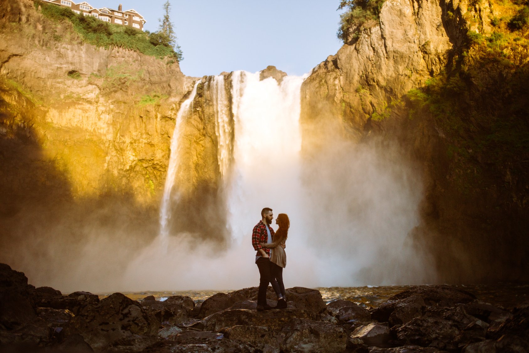 A couple stands close together in front of a large waterfall with mist rising from the rocks at the base, and a house visible atop the cliffs in the background.