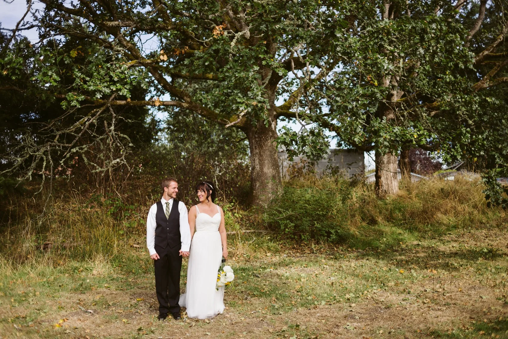 A bride and groom holding hands and walking outdoors on a grassy area under a large tree, smiling at each other. The groom is wearing a black vest, white shirt, and striped tie, while the bride is in a white wedding dress holding a bouquet of flowers