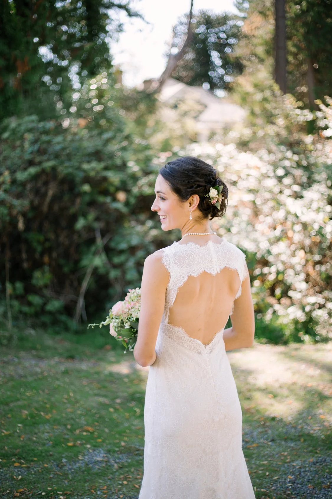 A bride in a white lace wedding dress with an open back, holding a bouquet of pink and white flowers, standing outdoors with greenery and trees in the background.