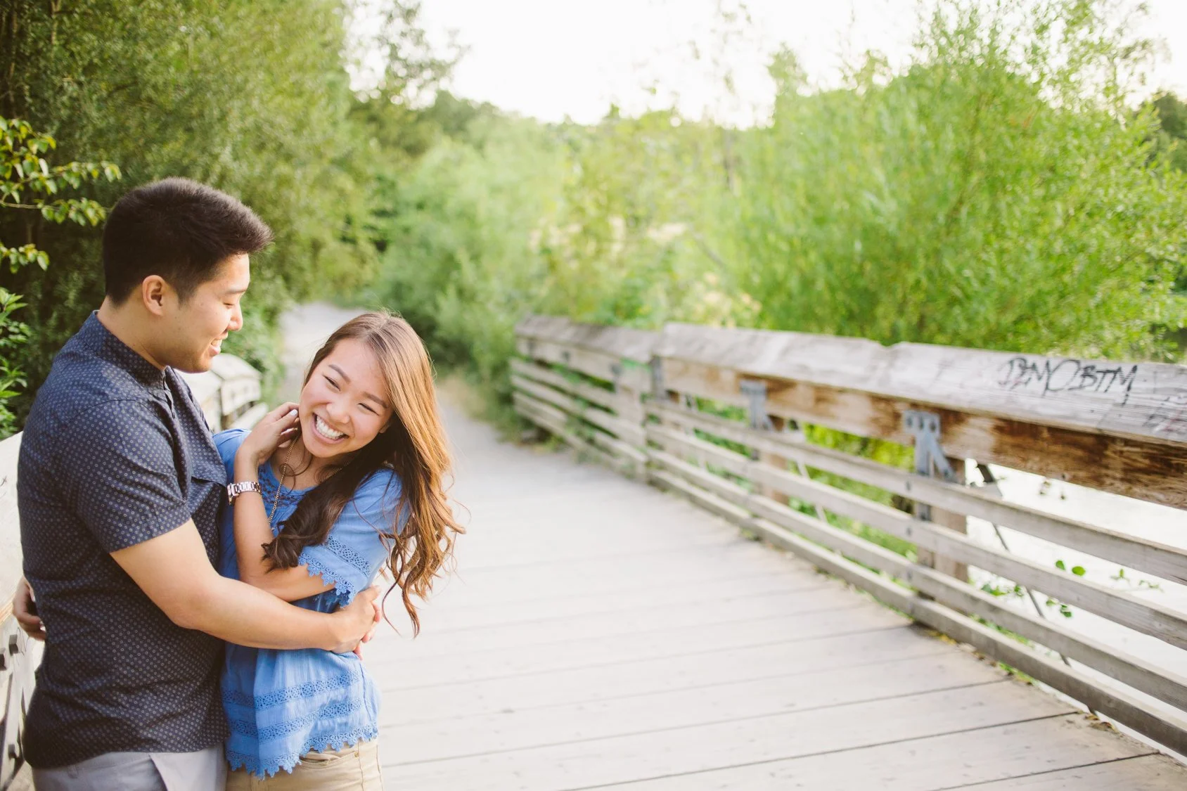 A young man and woman sharing a joyful moment on a wooden bridge surrounded by green trees and foliage.
