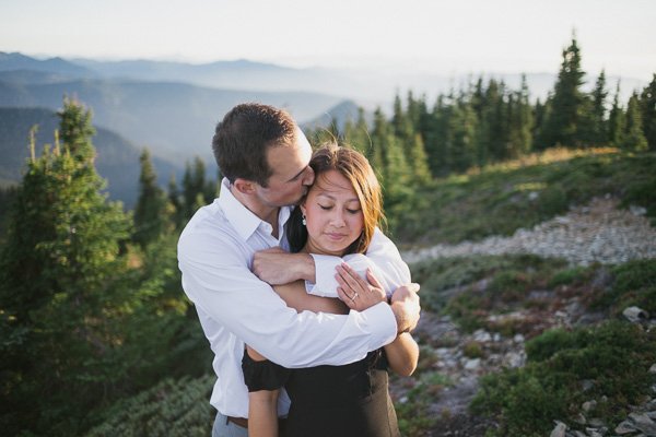 A man and woman embracing outdoors in a mountainous forested area, with trees and mountains in the background.