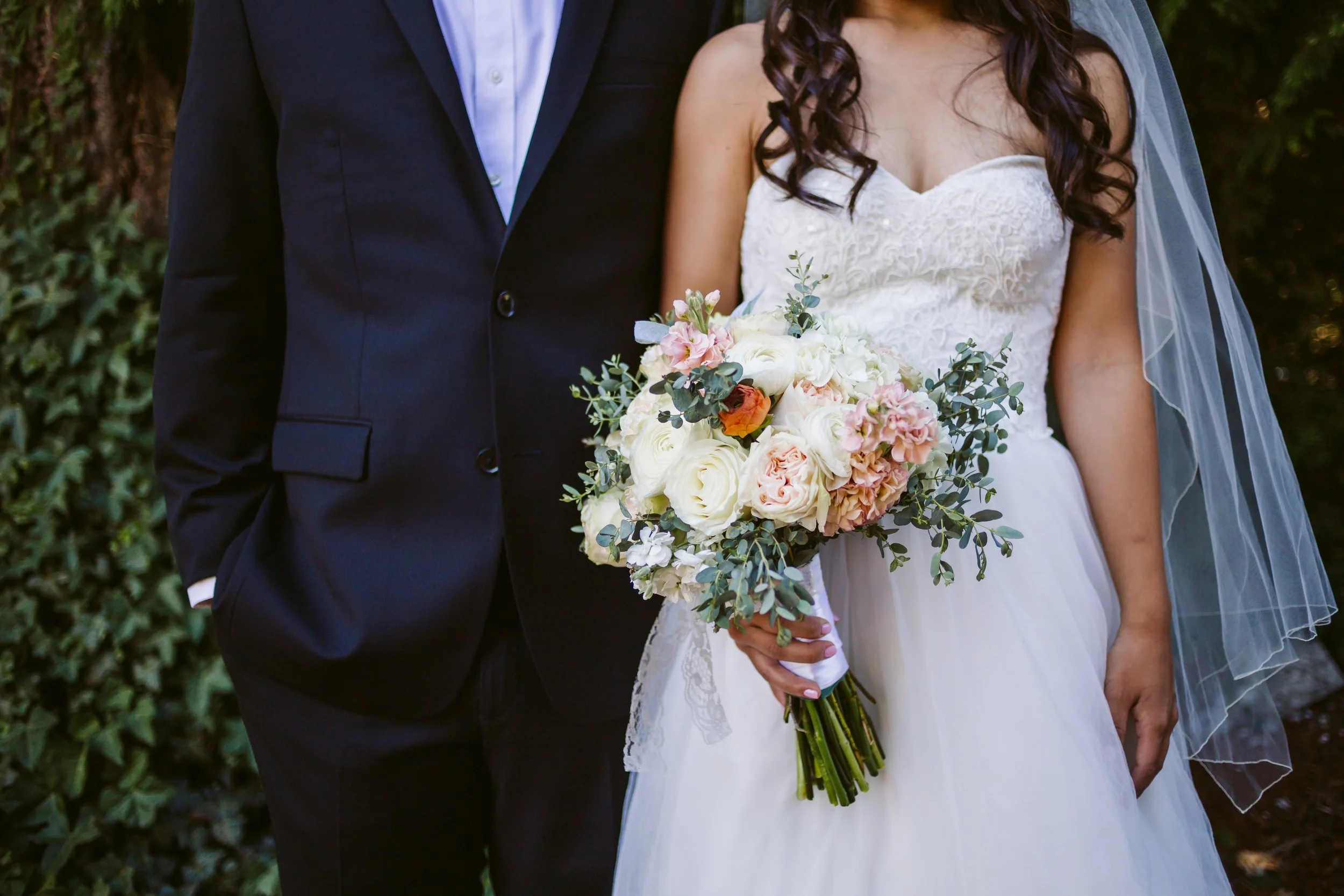 A bride in a white wedding dress holding a bouquet of pink and white flowers, standing next to a groom in a black suit, outdoors with greenery in the background.