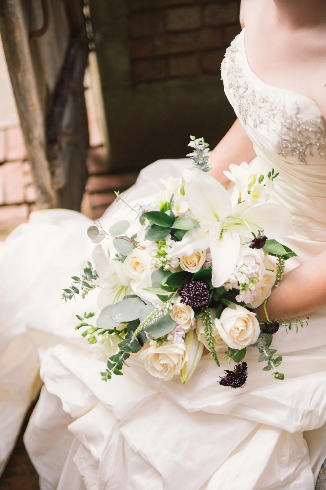 A bride in a white wedding gown holding a bouquet of white and cream flowers with green leaves and dark accents.