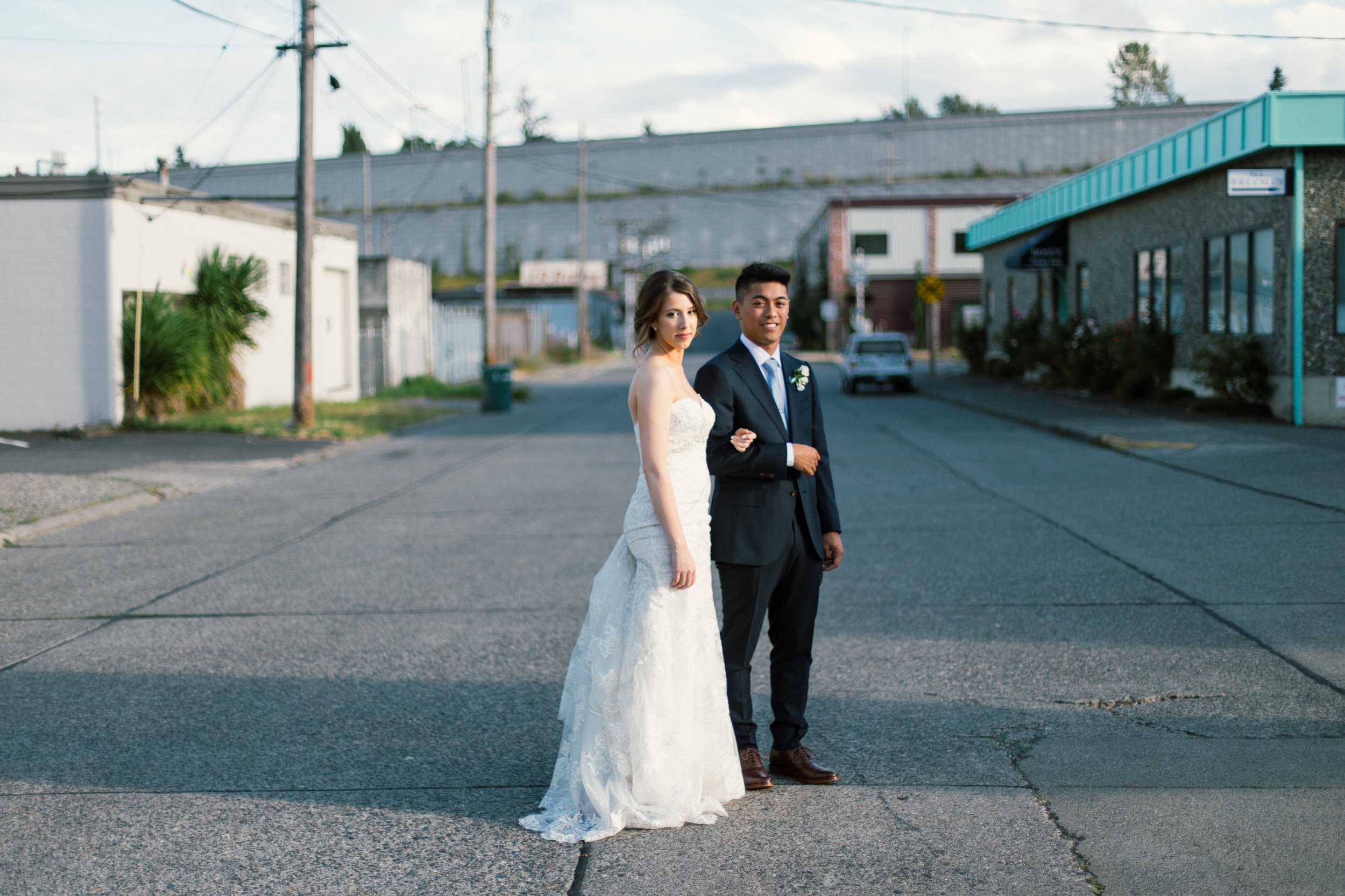 A bride and groom standing on a street in a small town, dressed in wedding attire, with the bride in a white gown and the groom in a dark suit. They are smiling and appear happy.