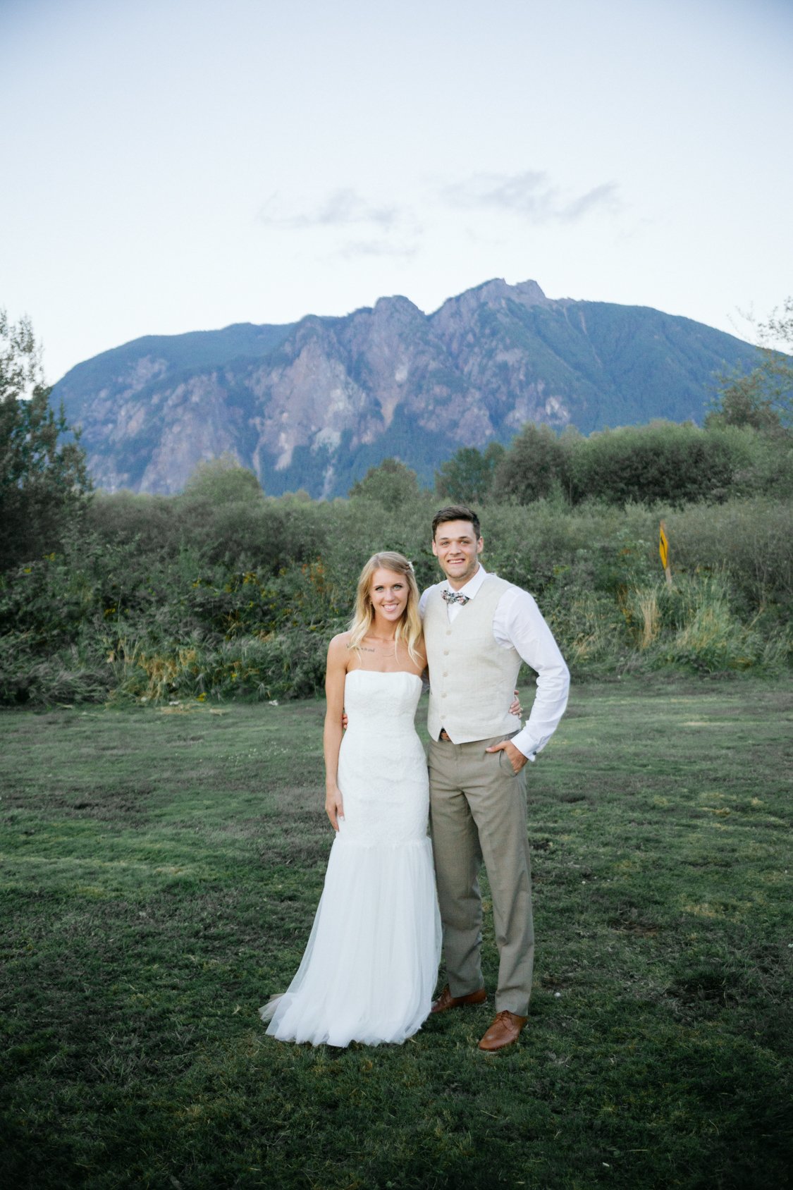 A bride and groom standing together outdoors in front of mountainous landscape. The bride is wearing a white strapless wedding dress, and the groom is wearing a white shirt, beige vest, and beige pants.