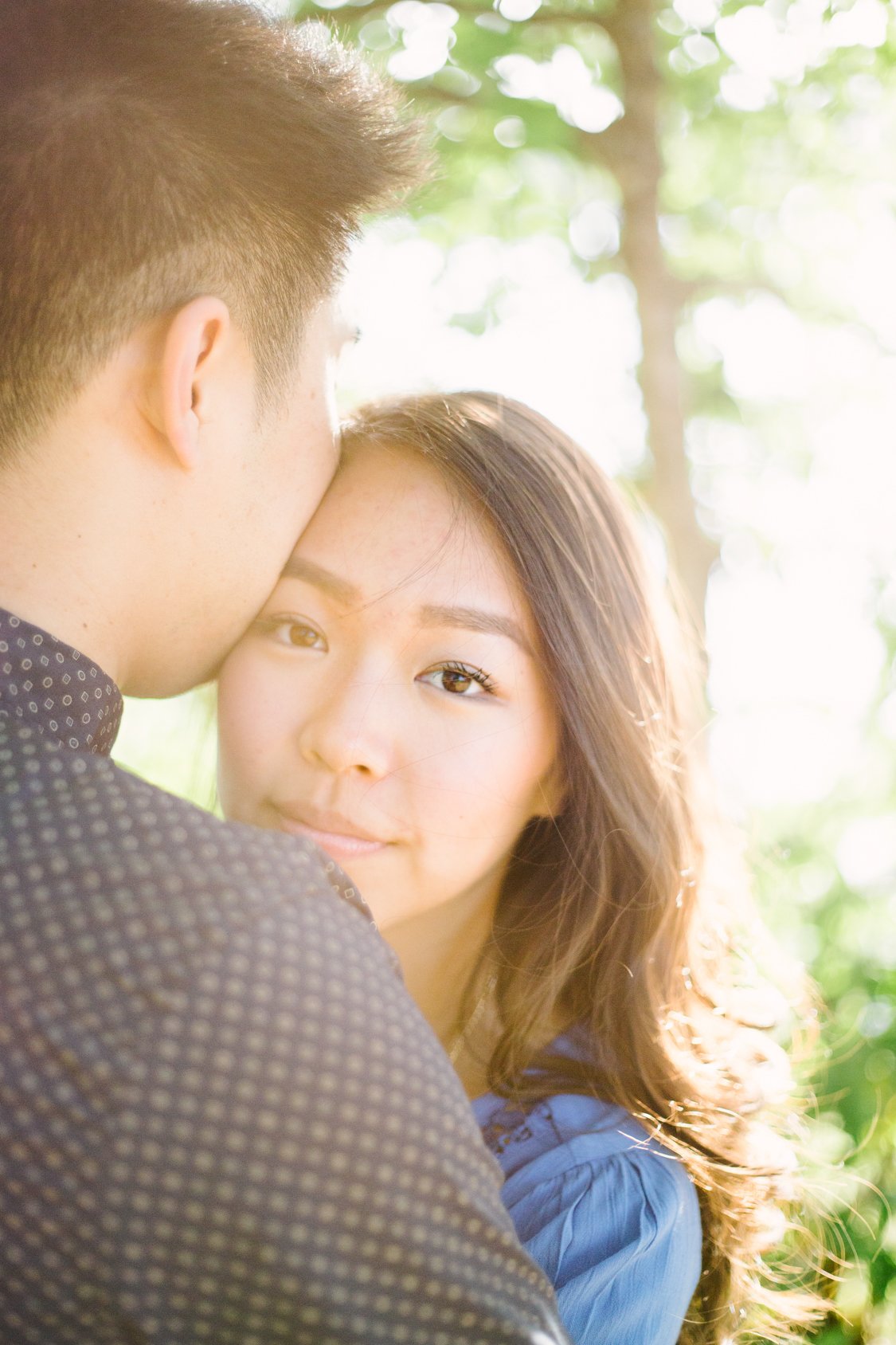 A close-up of a man kissing a woman on the forehead outdoors, with sunlight and green trees in the background.