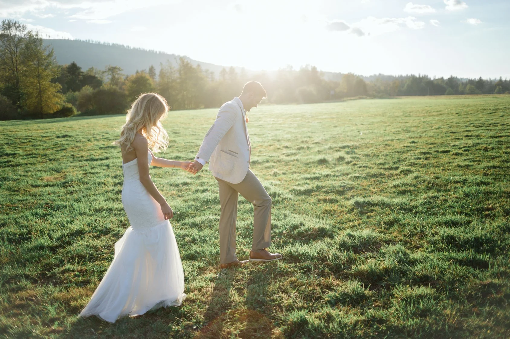 A bride and groom holding hands and walking in a grassy field during sunset or sunrise.