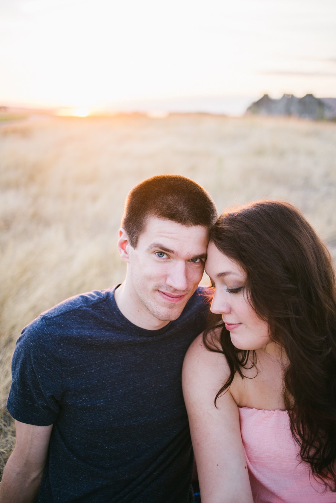 A young couple sitting close together outdoors during sunset, with the man smiling at the camera and the woman with her eyes closed leaning into him.