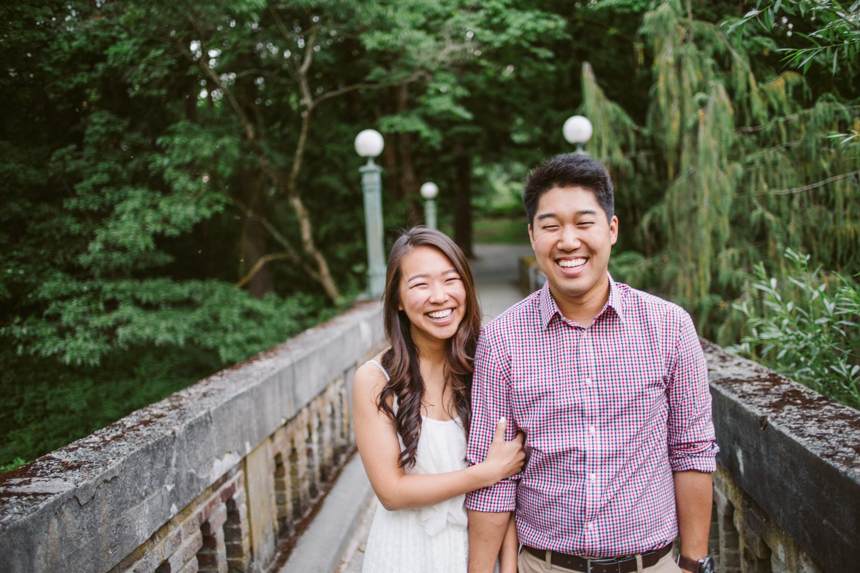 A happy couple standing on a stone bridge in a park, smiling and laughing, surrounded by green trees and bushes.