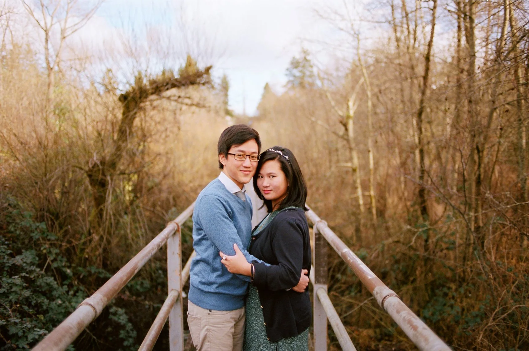 A couple hugging on a bridge surrounded by leafless trees in a park during autumn or winter.