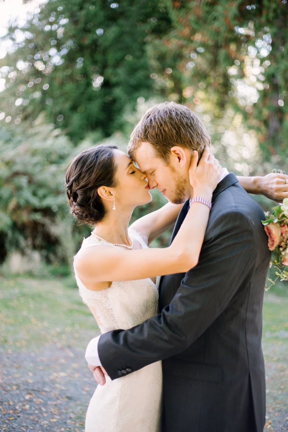 A bride and groom holding each other closely, with their foreheads touching, outdoors in a garden or park, during their wedding, with greenery and trees in the background.