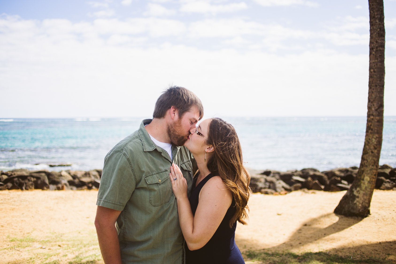 A couple sharing a kiss on a beach, with the ocean and a tree in the background.