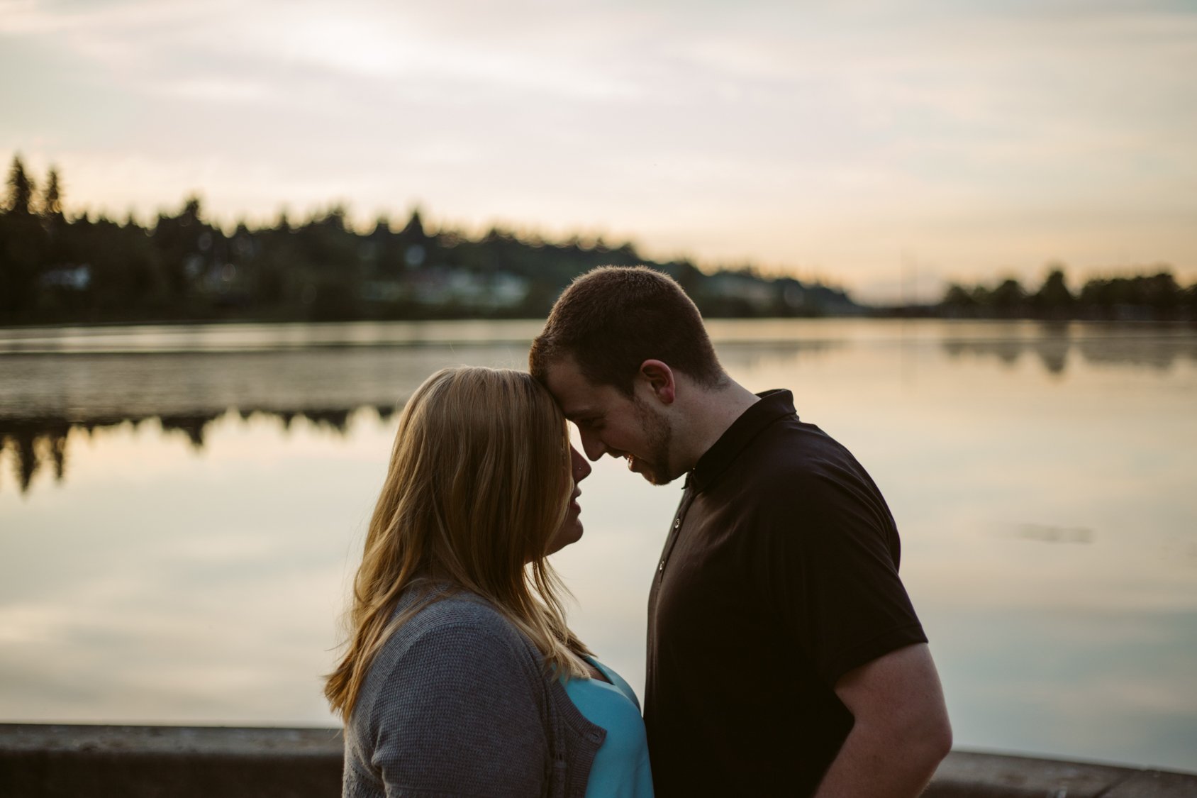 A couple standing close together by a body of water during sunset, with their foreheads touching and smiling.