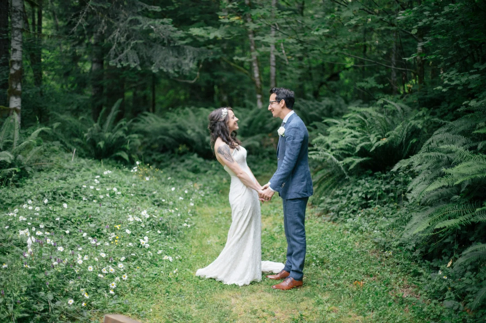 A bride and groom standing hand in hand in a lush green forest clearing, smiling at each other, with the bride in a white dress and the groom in a blue suit.