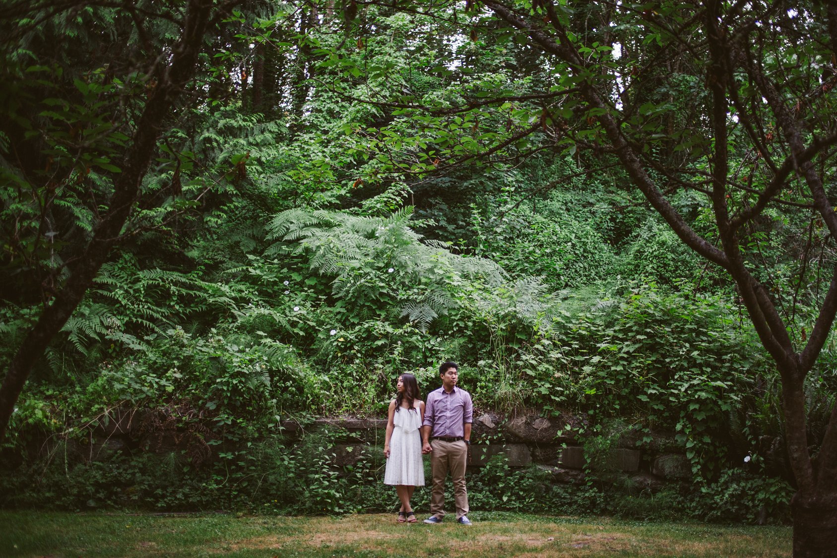 A couple stands holding hands in a lush, green forest near a stream, surrounded by dense foliage and trees.