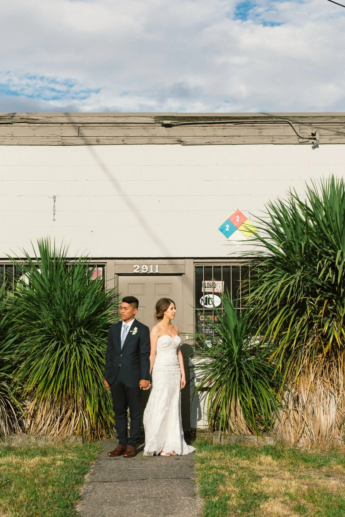 A couple dressed in wedding attire standing hand in hand in front of a building with the number 2911. The man is wearing a dark suit and the woman is wearing a strapless white wedding dress. Large green plants are on either side of them, and a climat