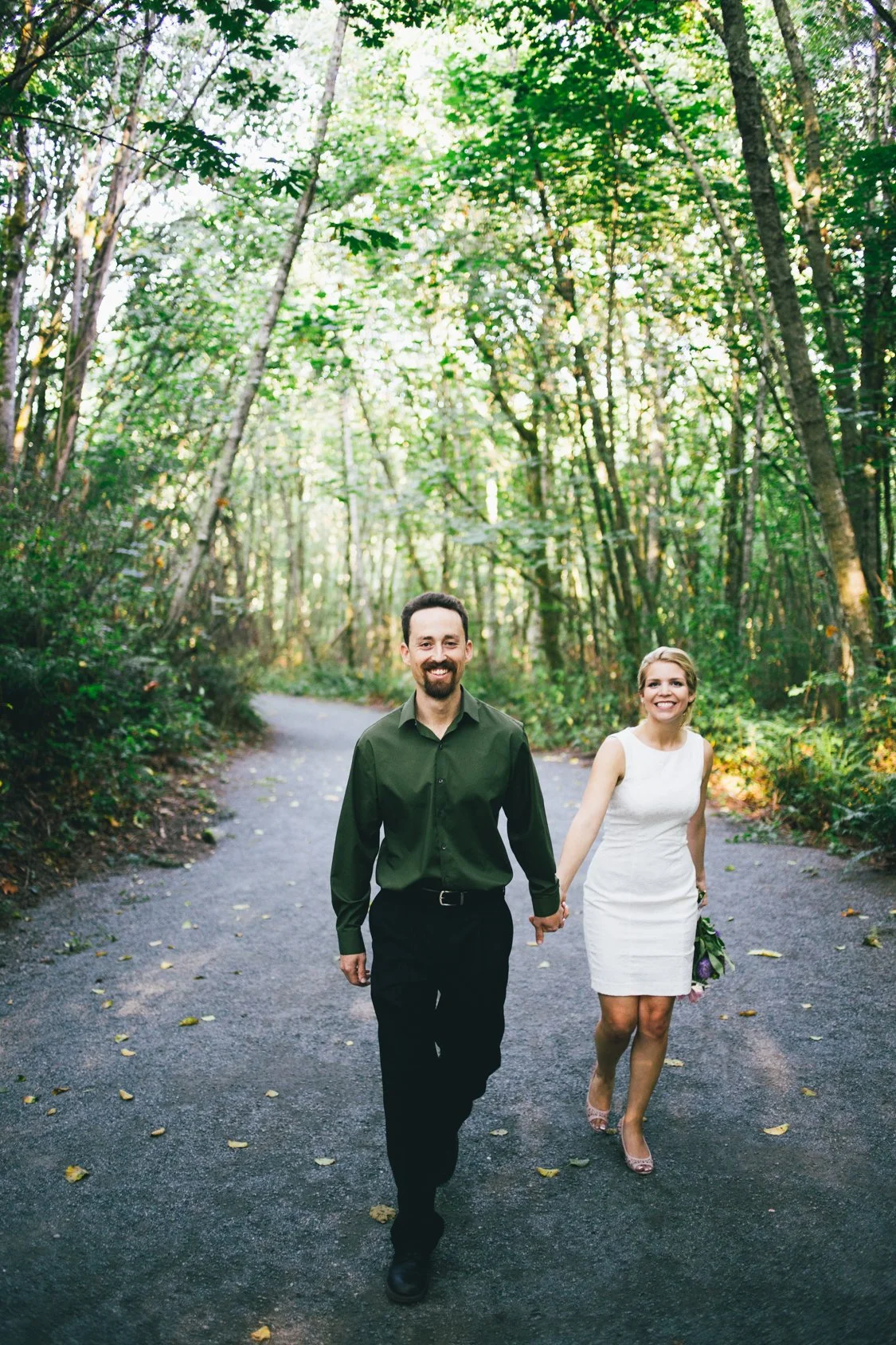 A smiling man and woman holding hands and walking on a forest path surrounded by green trees, with sunlight filtering through the leaves.