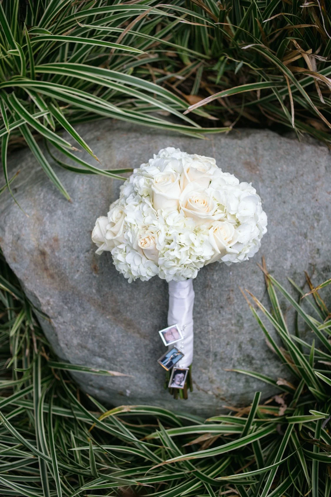 Wedding bouquet with white roses and hydrangeas resting on a gray stone, surrounded by green grass with variegated leaves. Small photographs are attached to the handle of the bouquet.