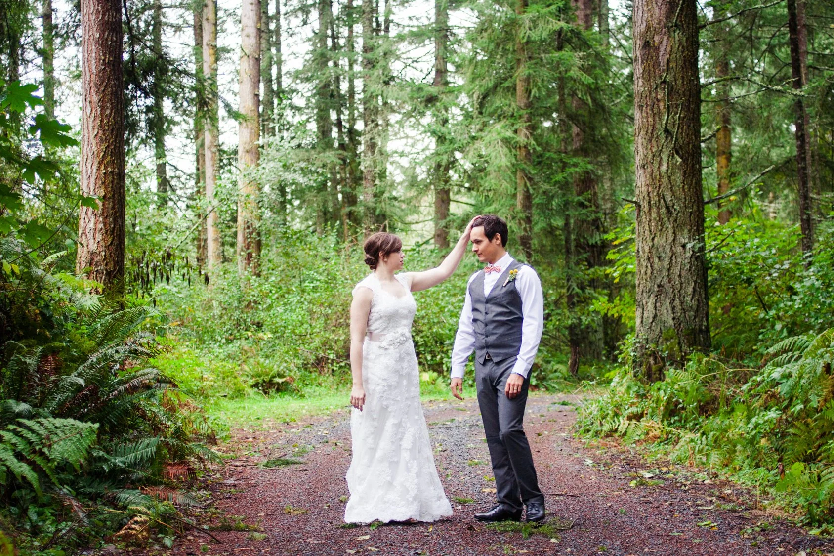 A bride in a white lace wedding dress and a groom in a gray vest and pants stand in a lush green forest, with the bride gently touching the groom's head.