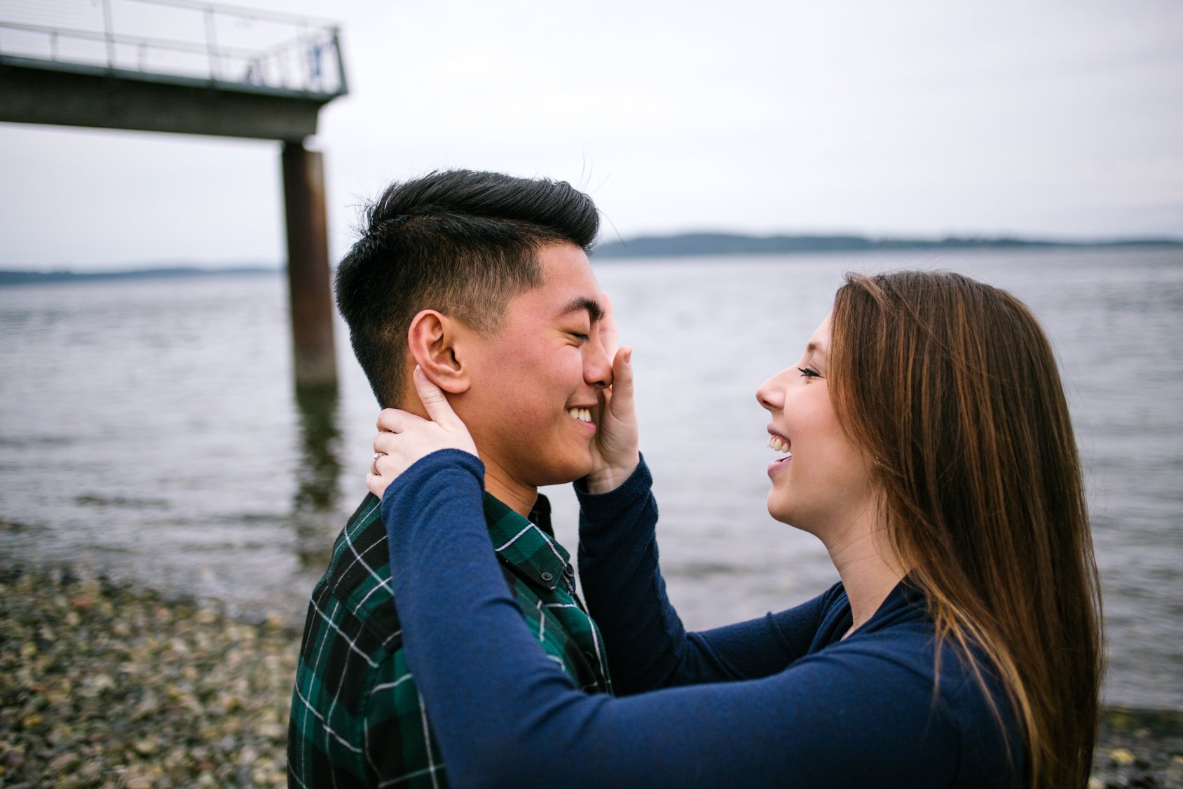 A young couple smiling and touching each other's faces near a body of water under a cloudy sky, with a pier in the background.