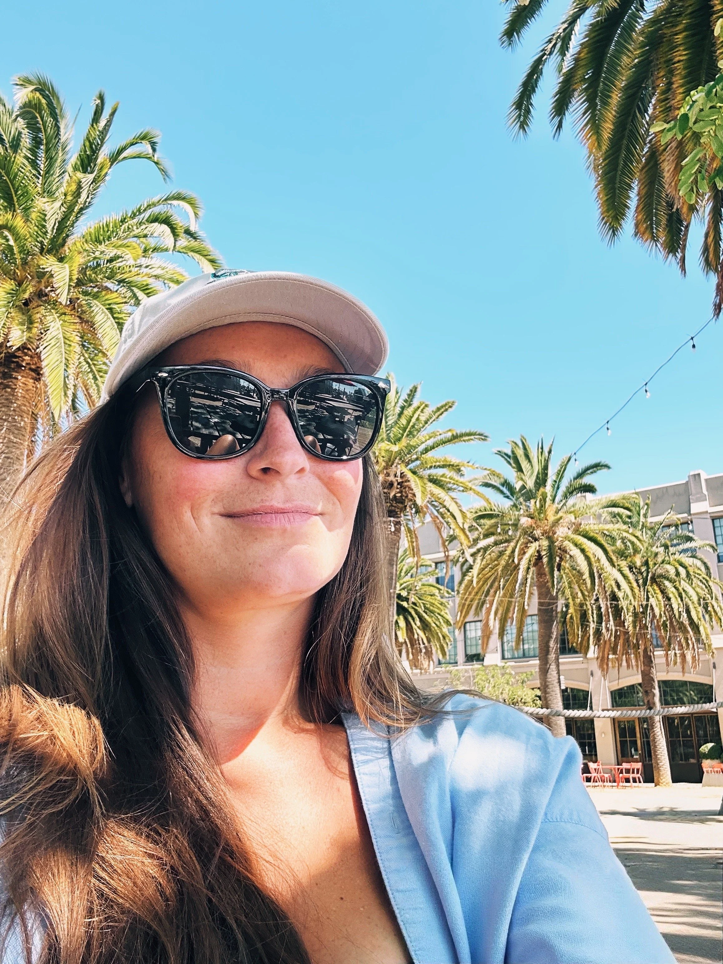 A smiling woman wearing sunglasses and a white cap, outdoors on a sunny day, with palm trees and a building in the background.