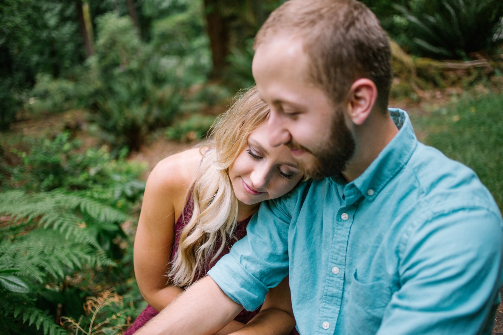 A couple sitting closely together outdoors with their heads touching and eyes closed, surrounded by green foliage.