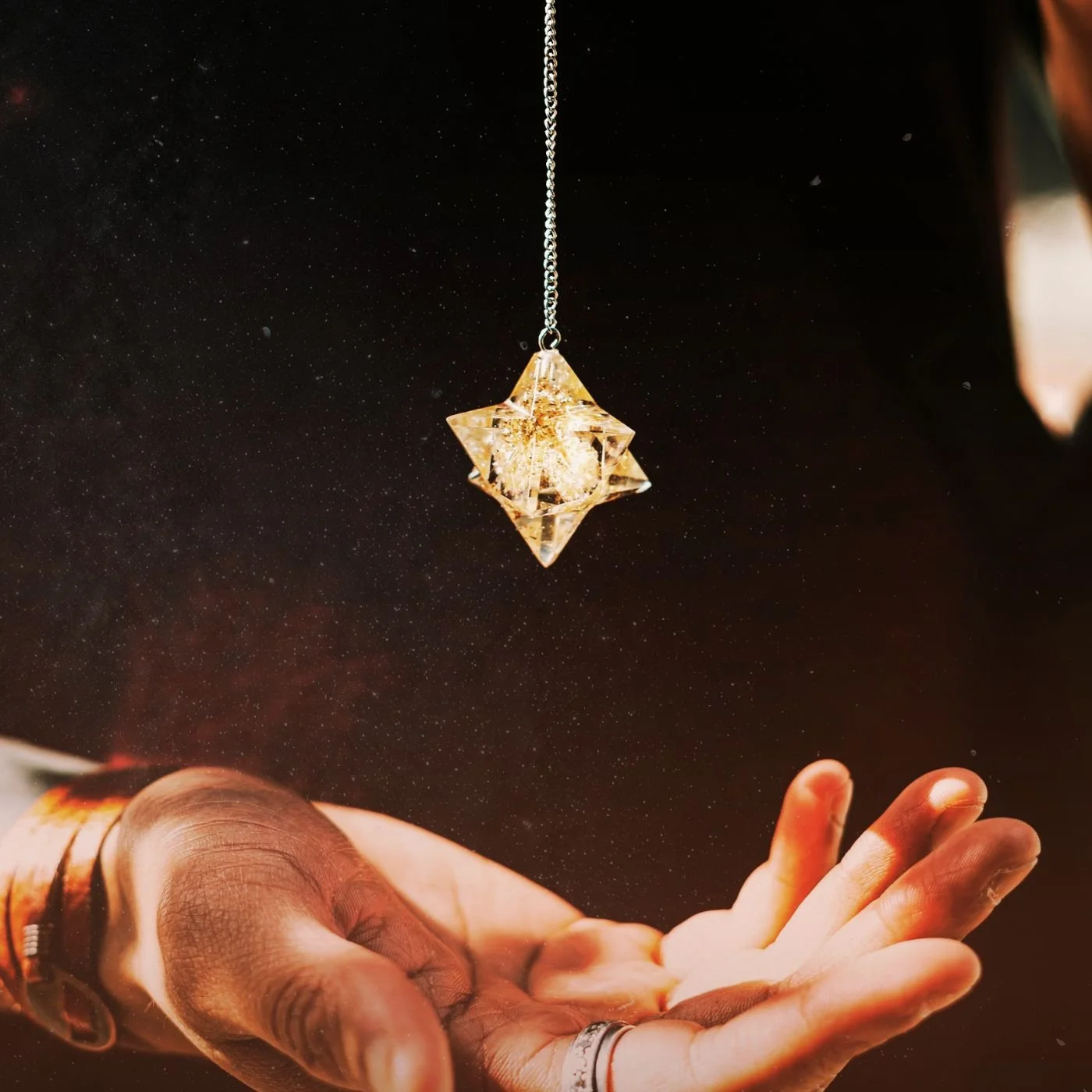 A person holding out their hand with fingers slightly apart, underneath a floating geometric pendant with a chain, against a dark, starry background.