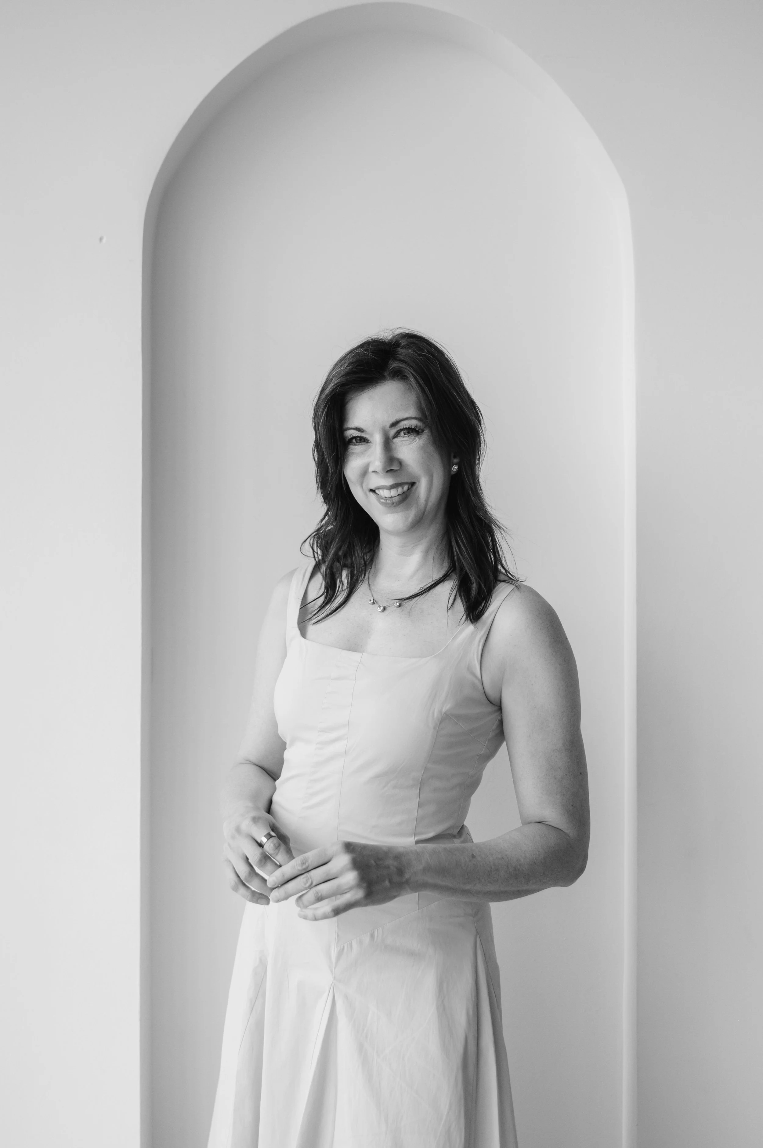 Black and white photo of a woman with dark hair wearing a light-colored dress, smiling, standing against a plain wall with an arched niche.