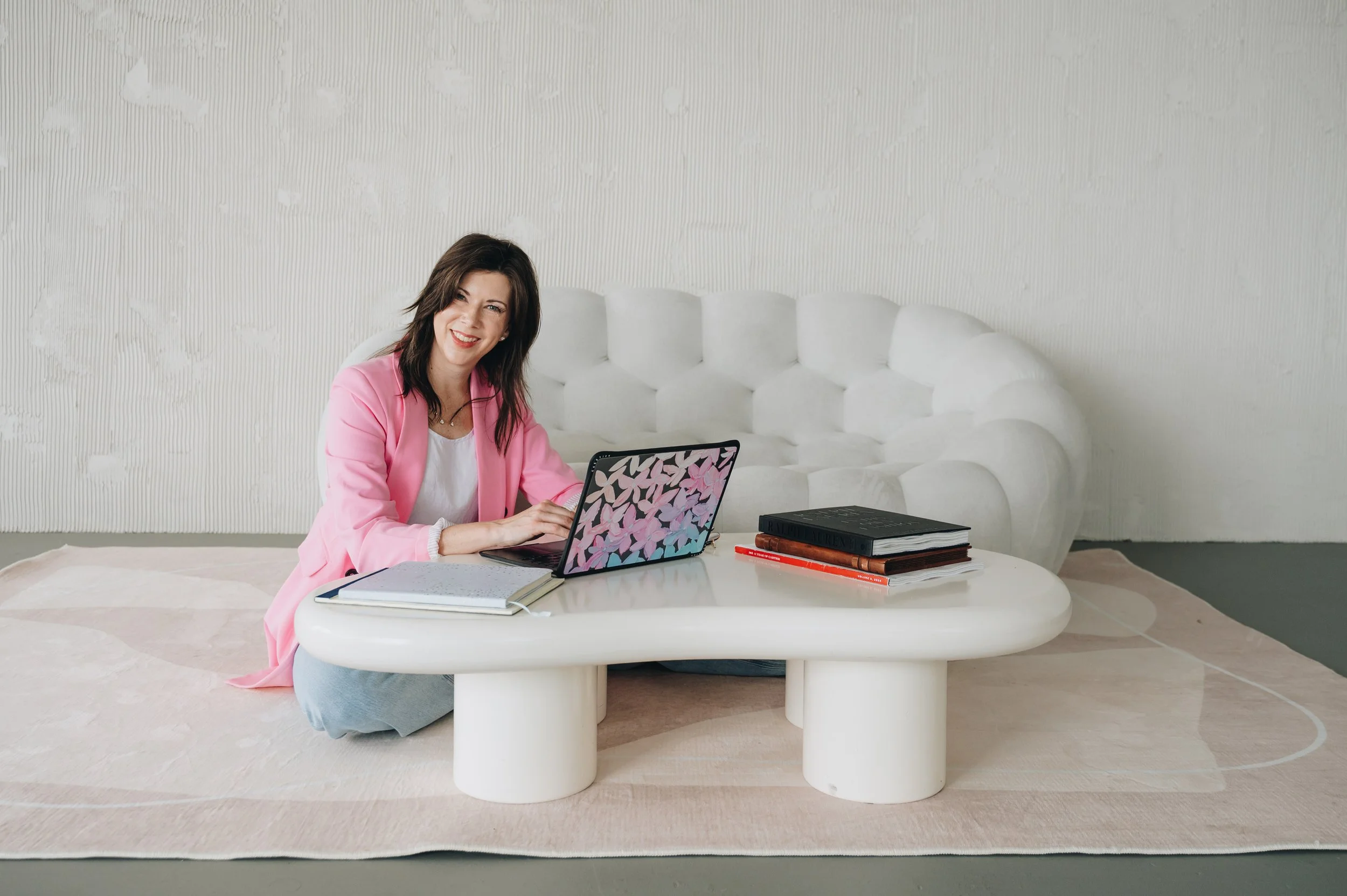 A woman with dark hair smiling and sitting on the floor at a white, round table with a laptop, notebooks, and books in a minimalistic room with a textured white wall and a white plush sofa in the background.