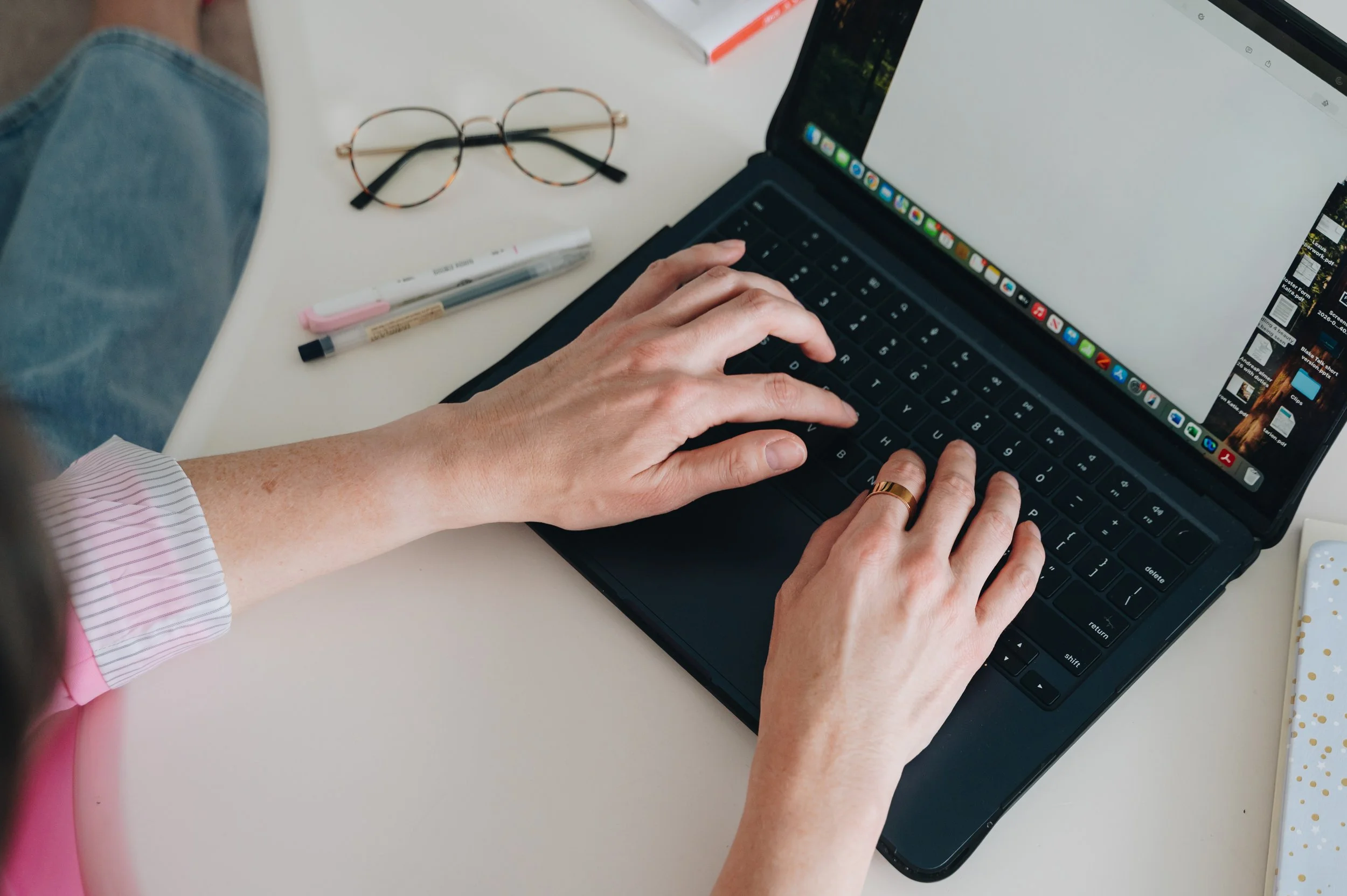 A person typing on a laptop keyboard at a white desk, with a pair of glasses, a marker, and a closed book nearby.