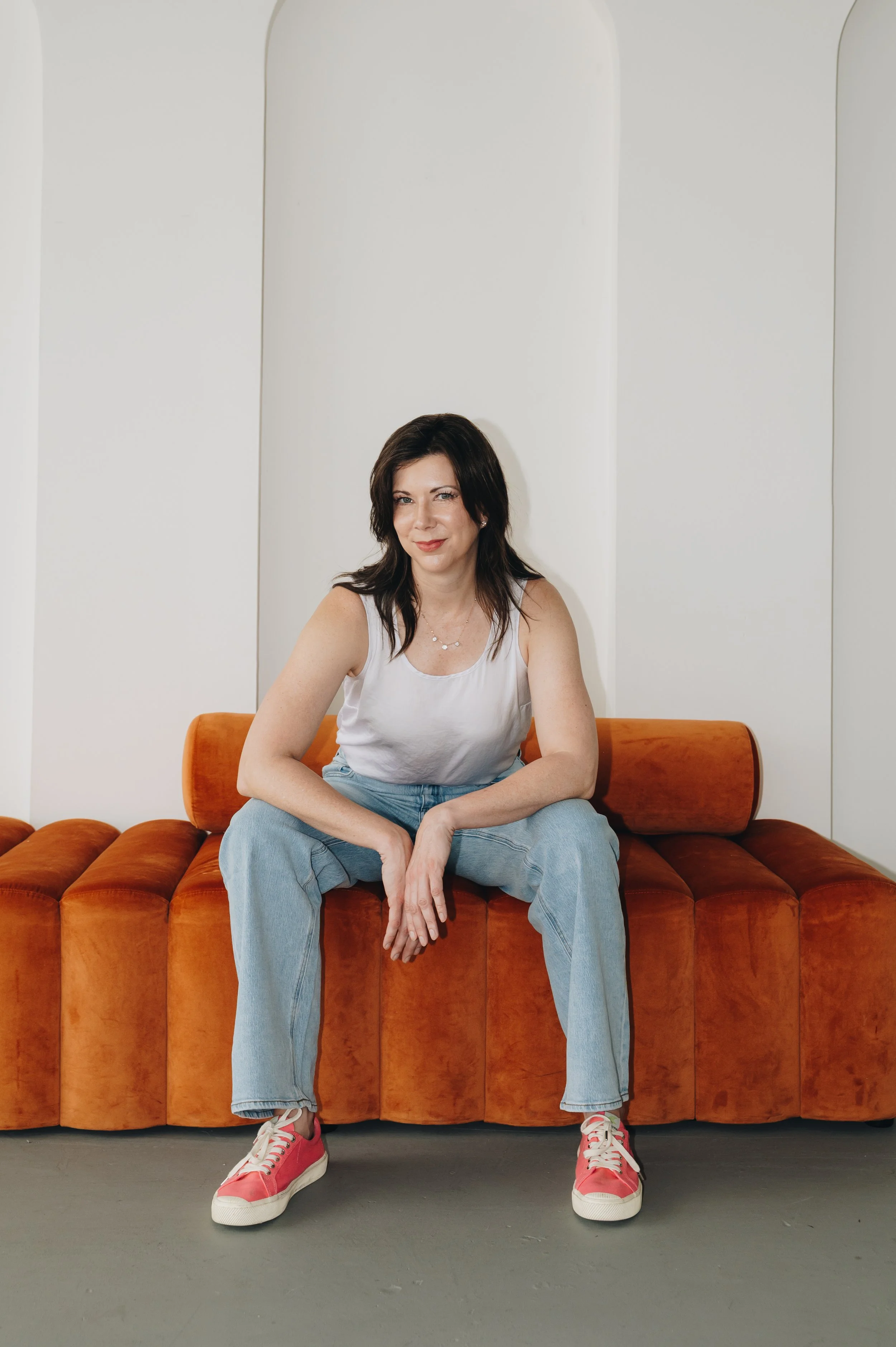 A woman with black hair, wearing a white tank top, light blue jeans, and pink sneakers, sitting on an orange velvet sofa with her legs spread apart, in front of a white wall with curved panels.