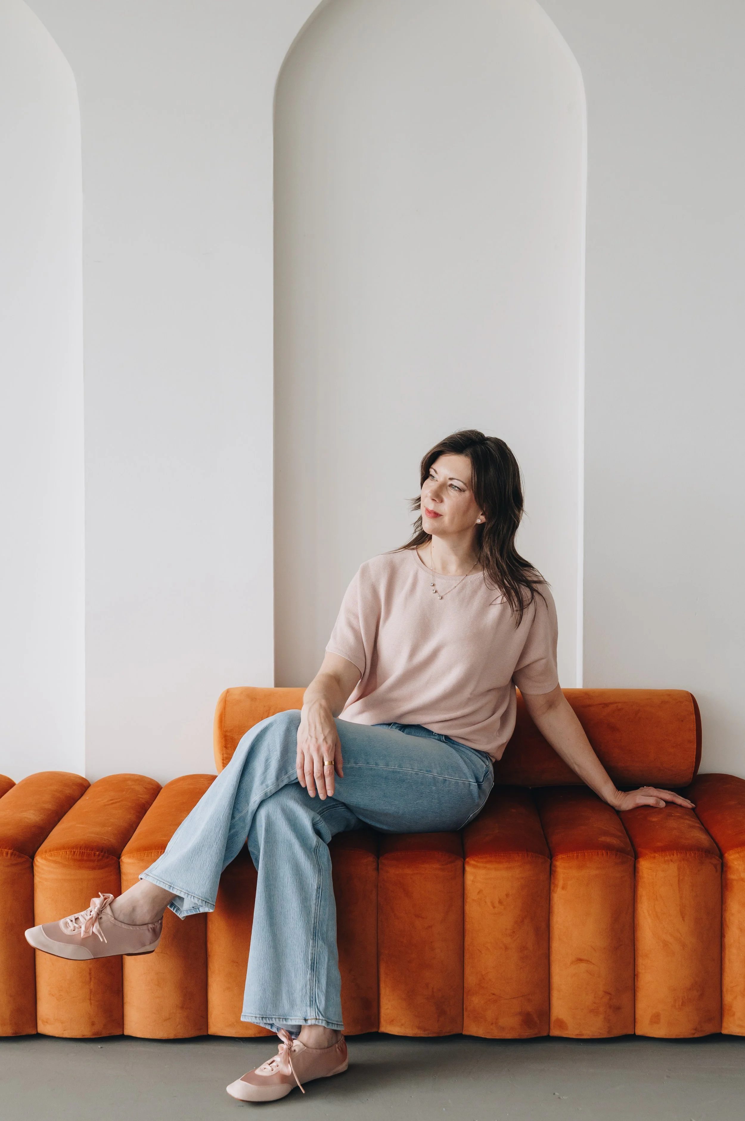 A woman sitting on an orange velvet tufted sofa in a room with white arched walls. She wears a light pink T-shirt, light blue jeans, and pink sneakers.
