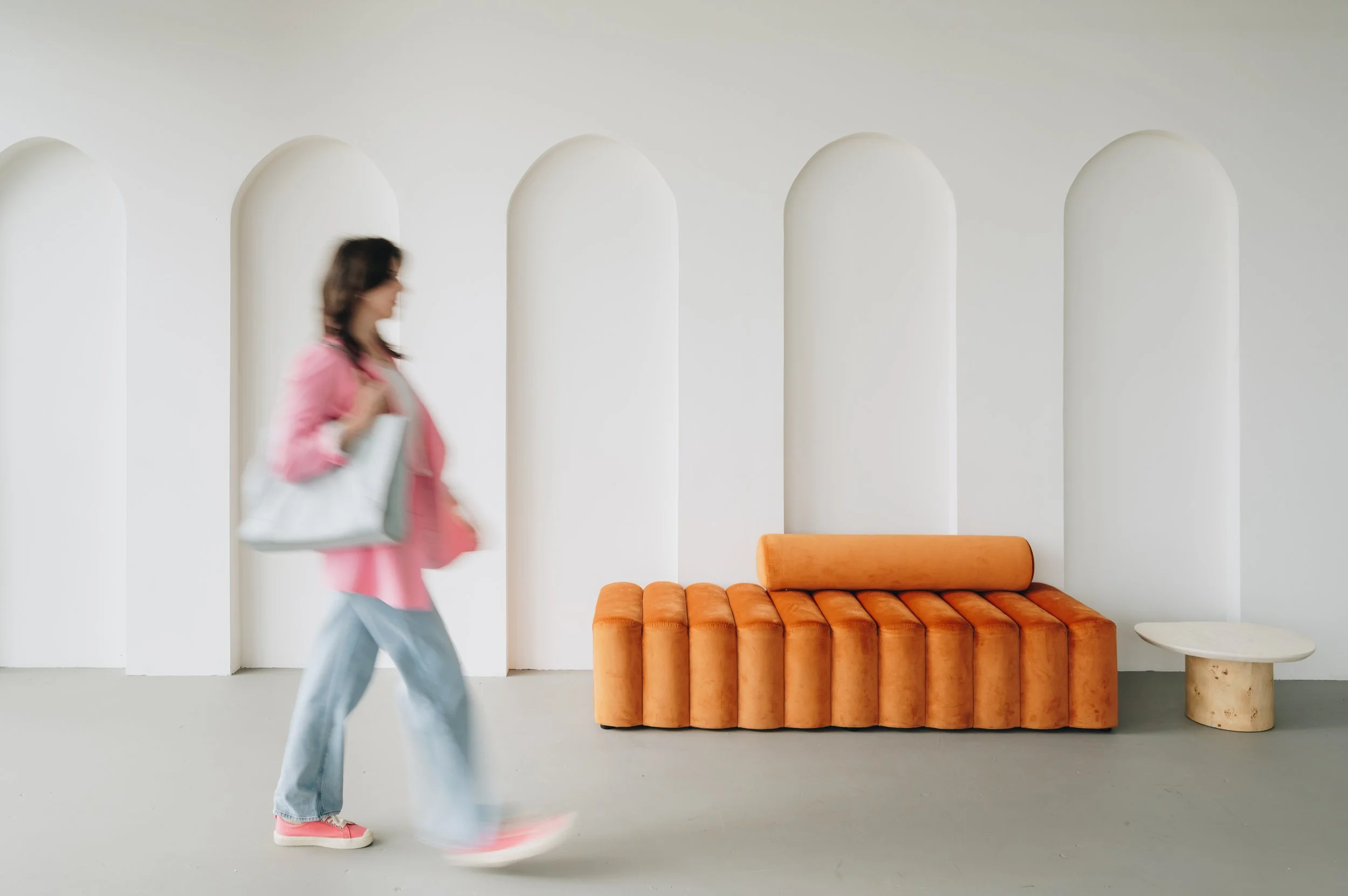 A woman walking past modern furniture against a white wall with arched niches, including an orange sofa and marble side table.